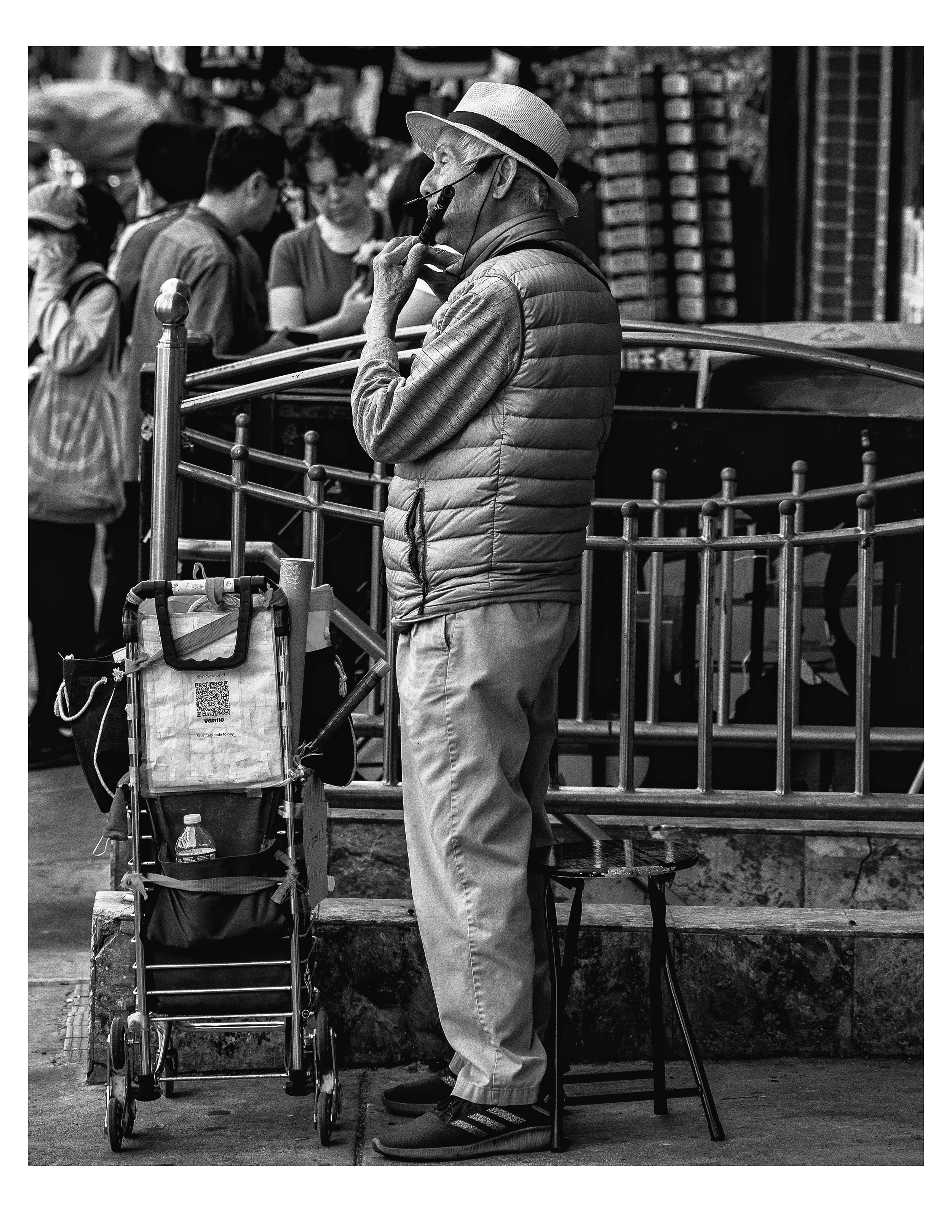 An elderly man with a hat, headphones, and a puffer vest stands next to a shopping cart, deep in thought, with multiple people blurred in the background at an outdoor market or street scene.