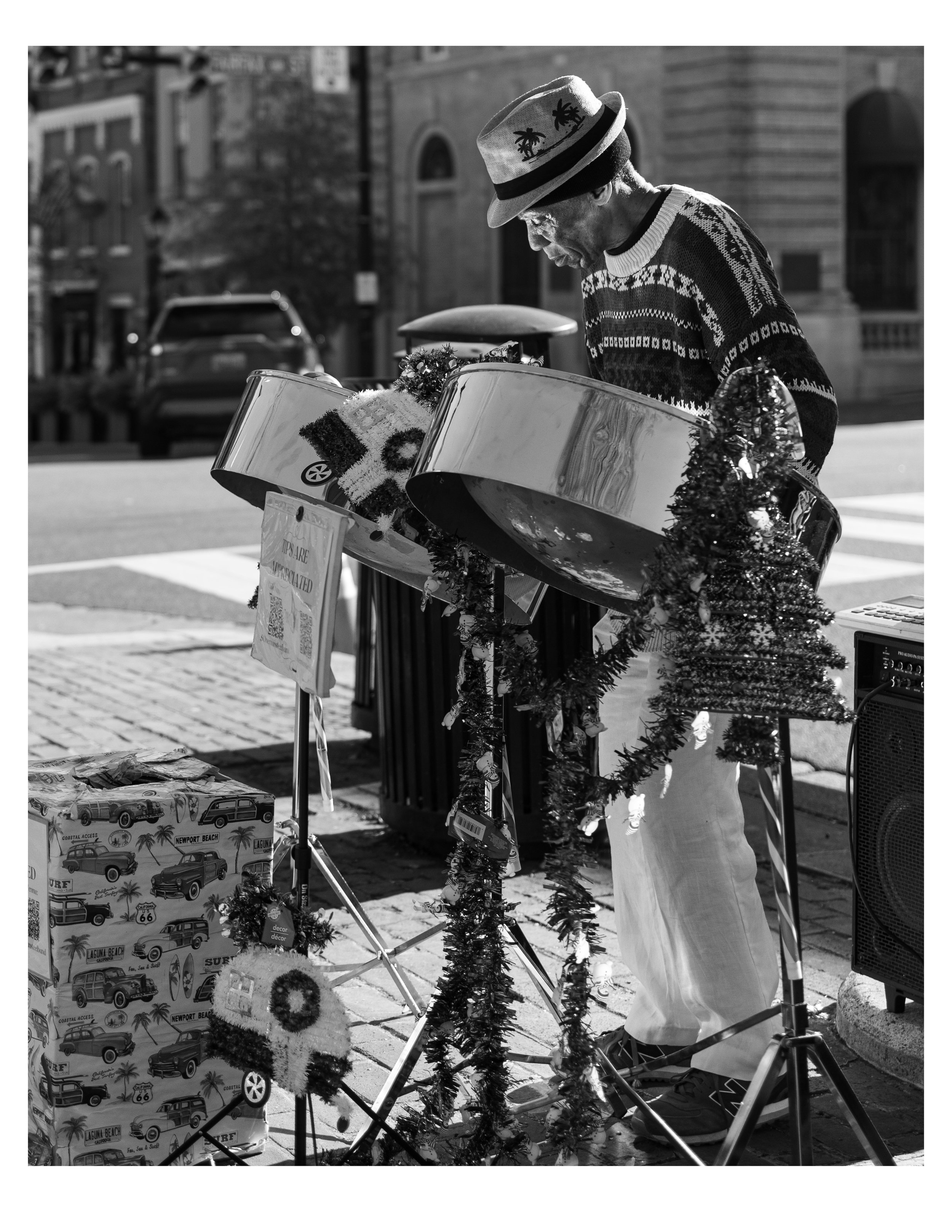 A man wearing a hat and Christmas sweater playing steel drums on a city street decorated with garlands. There are boxes with surf-themed illustrations nearby and a small Christmas tree in front of the drums. The scene appears to be during the holiday