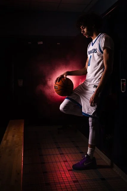 A young basketball player in a white jersey with blue accents and purple shoes stands in a dark locker room, holding a basketball with a red light behind him.