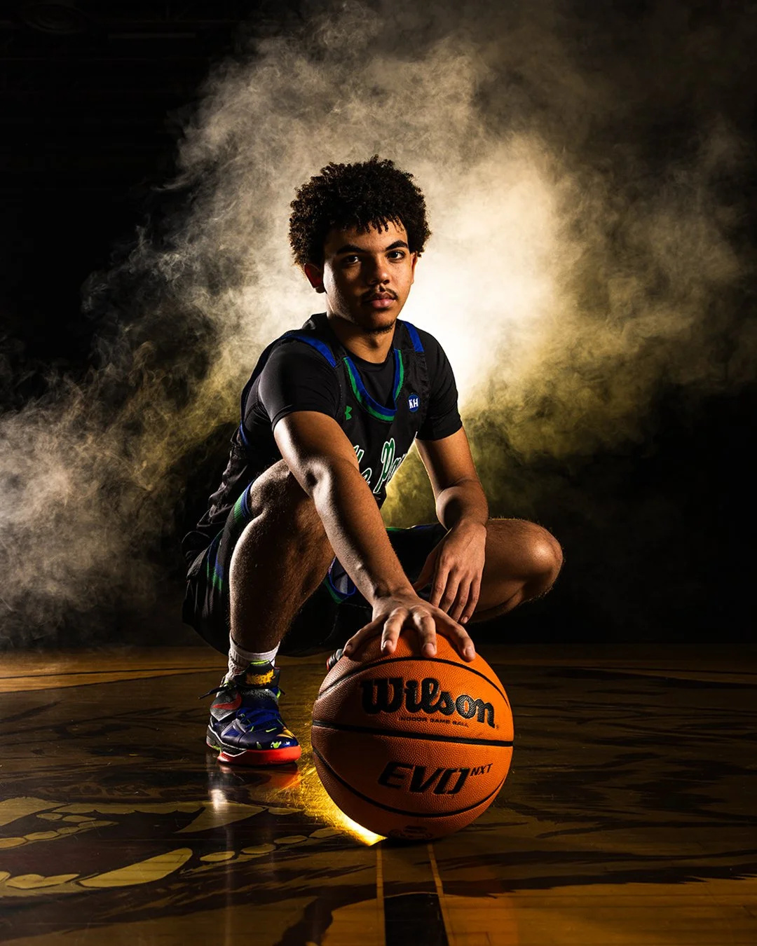 A young male basketball player crouched on the court with one hand on a Wilson EVO NXT basketball, surrounded by a smoky background with warm lighting.