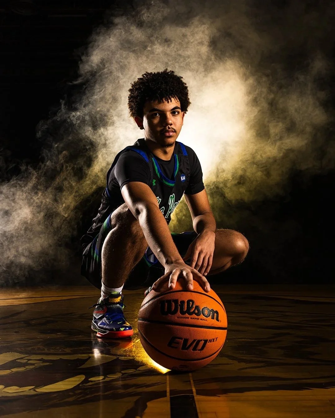A young male basketball player crouching on a basketball court, holding an orange Wilson basketball with his right hand. He is wearing sports attire including a jersey, shorts, and sneakers, with colorful lighting and a smoke-like background.