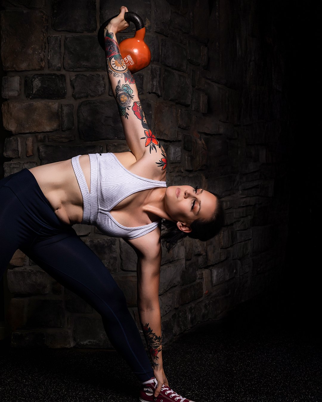 A woman with tattoos on her arm and leg wearing a white sports bra and black leggings, performing a yoga pose with her hand on the floor and holding an orange kettle bell overhead.