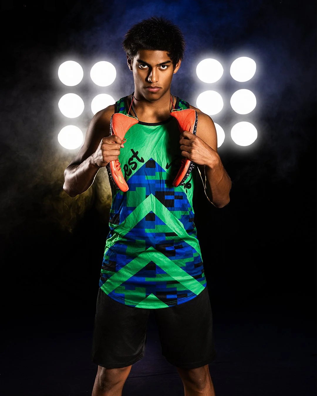 A young male athlete in colorful sportswear holding orange athletic shoes in front of bright stage lights.