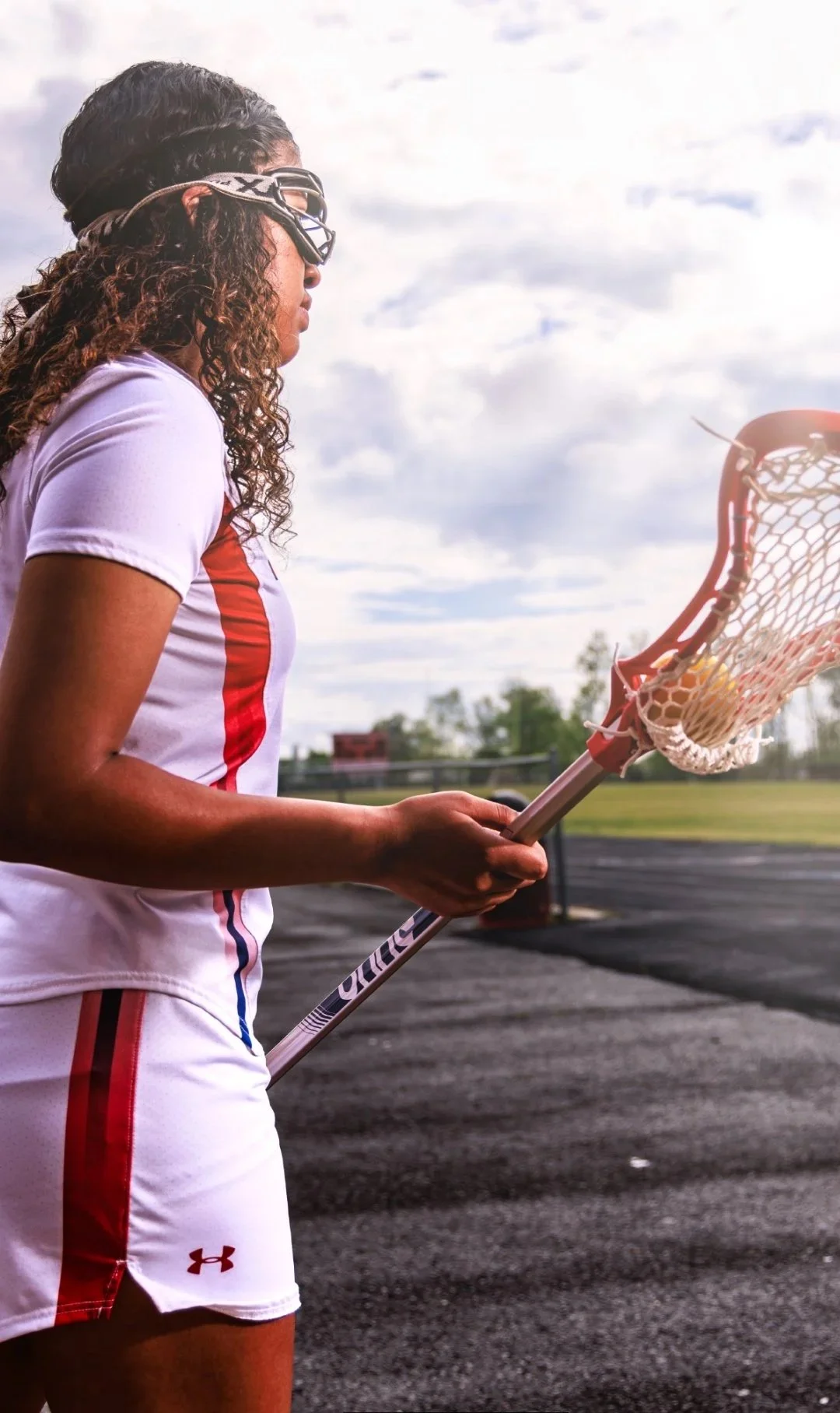 A female lacrosse player wearing protective goggles, a white and red uniform, holding a lacrosse stick, standing on an outdoor field with a cloudy sky in the background.