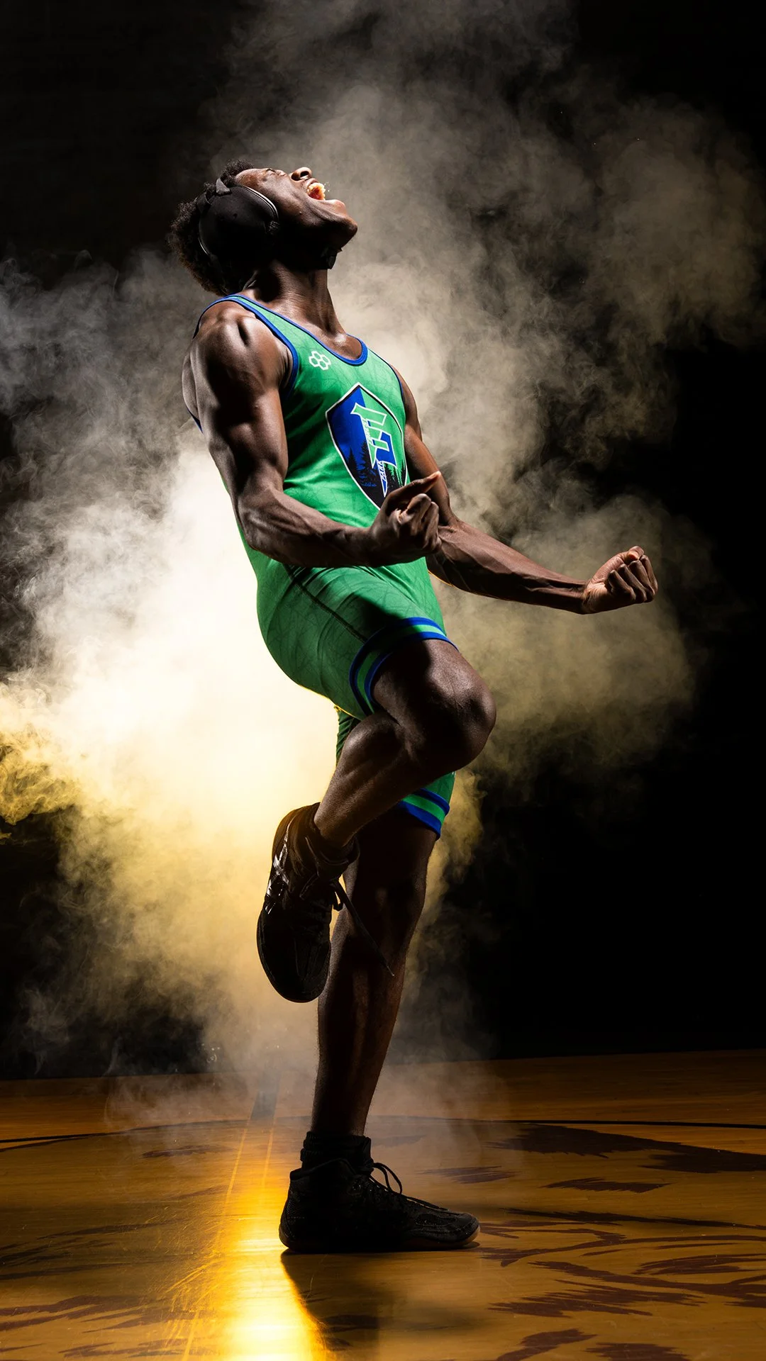 A muscular man in a green sports uniform with a logo, wearing headphones, shows a victorious pose with clenched fists and flexed muscles, standing on a wooden floor with smoke and dramatic lighting.