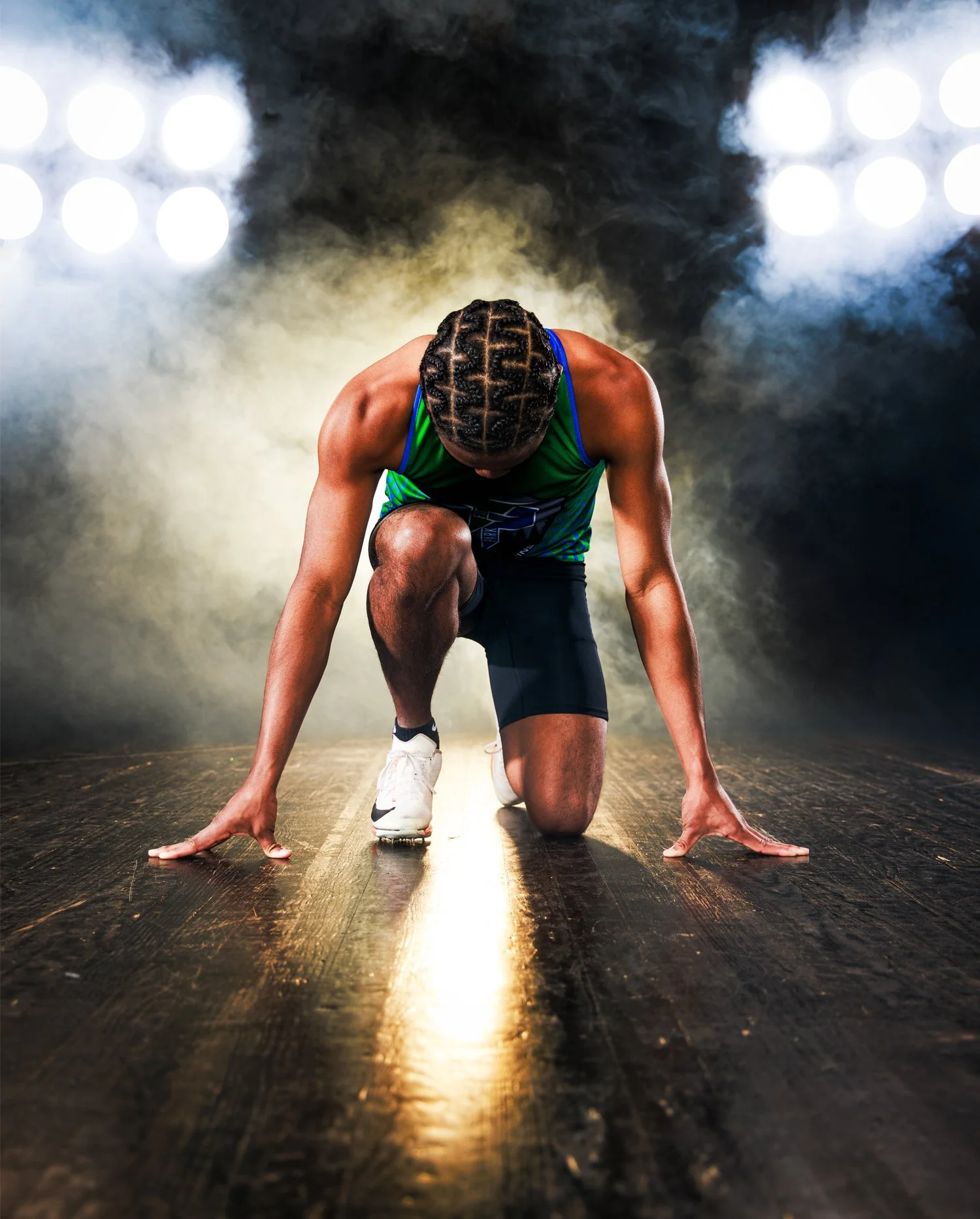 A male athlete in green sportswear, with braided hair, is kneeling on the floor in a starting position for a race on a wooden track, with bright stage lights and smoke in the background.