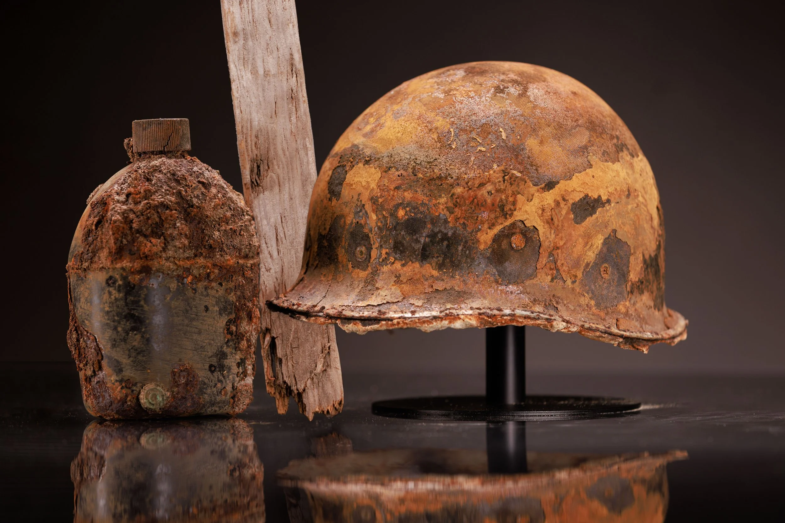 Photo of three rusty, aged helmets and a rusty bottle on a reflective black surface against black background.