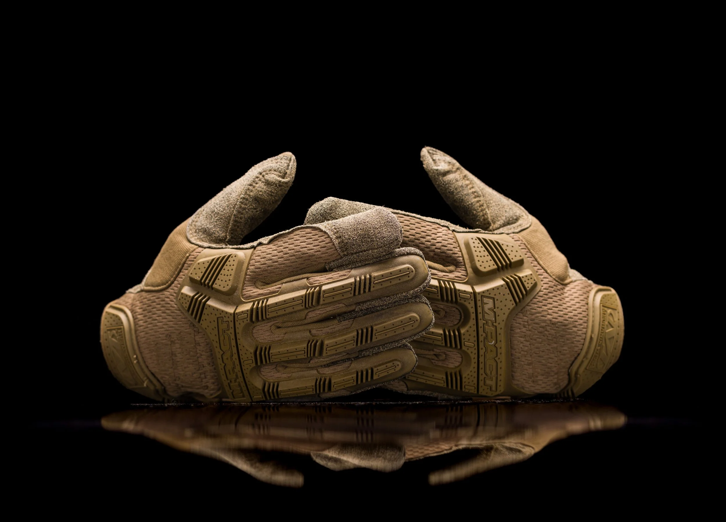 A close-up of a pair of tactical gloves with a rugged textured surface, held together with the fingers intertwined, against a black background.