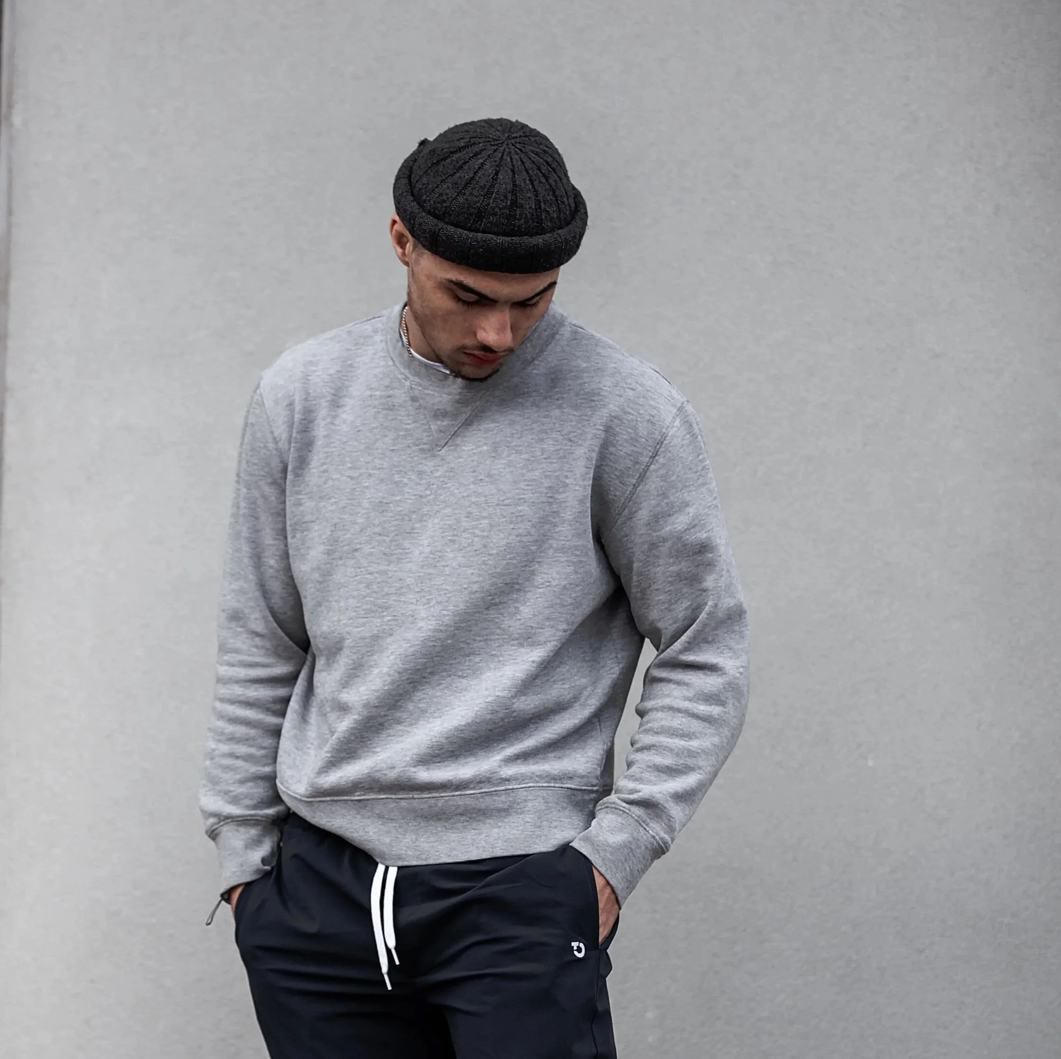 A young man is standing against a plain gray wall, wearing a dark knitted beanie, a light gray sweatshirt, and black track pants with white stripes, looking downward with a relaxed posture.