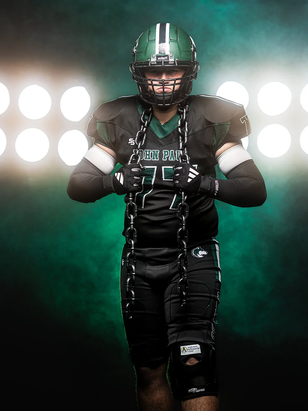 American football player in uniform, wearing a helmet and gloves, holding a chain with a determined expression, on a dark background with bright lights.