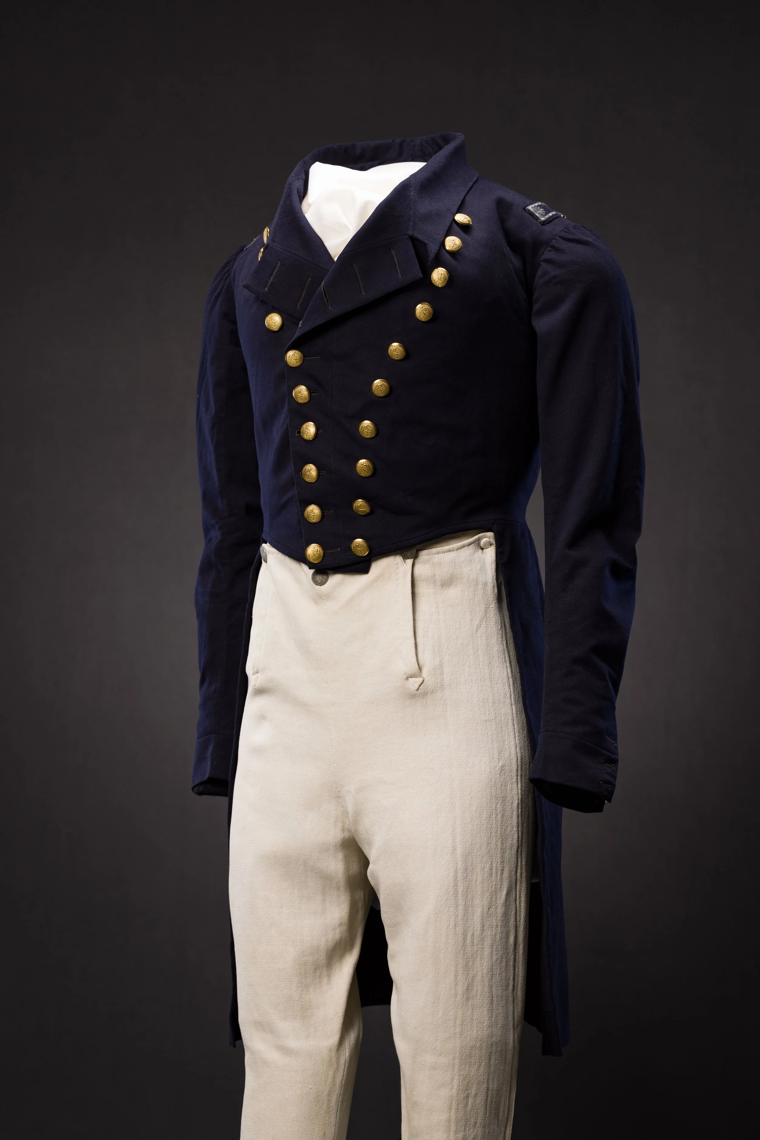 A vintage military uniform jacket with gold buttons, white shirt underneath, and beige trousers on display against a dark background.