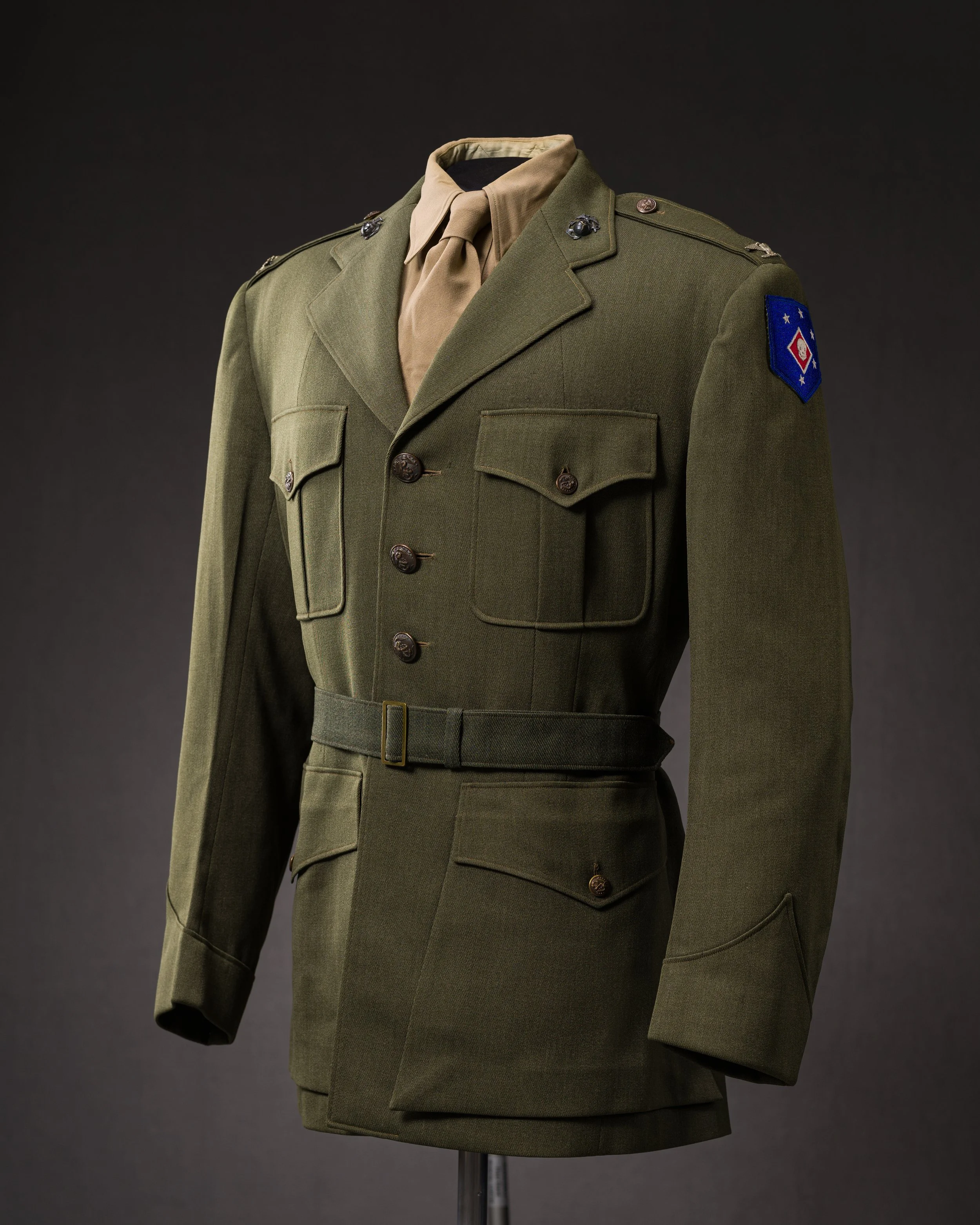 A military uniform on a mannequin, featuring an olive green jacket with gold buttons, a tan shirt with a matching tie, and a blue patch with white stars and a red emblem on the shoulder.