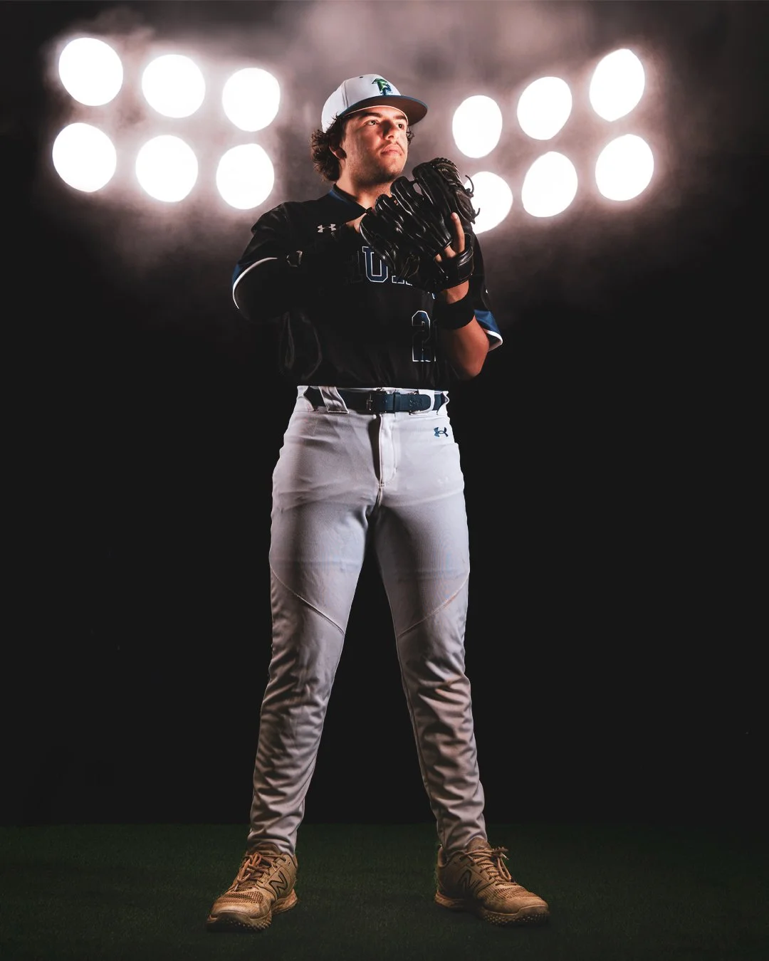 A male baseball player in uniform and a cap stands with hands together, holding a baseball glove, in front of bright stadium lights at night.