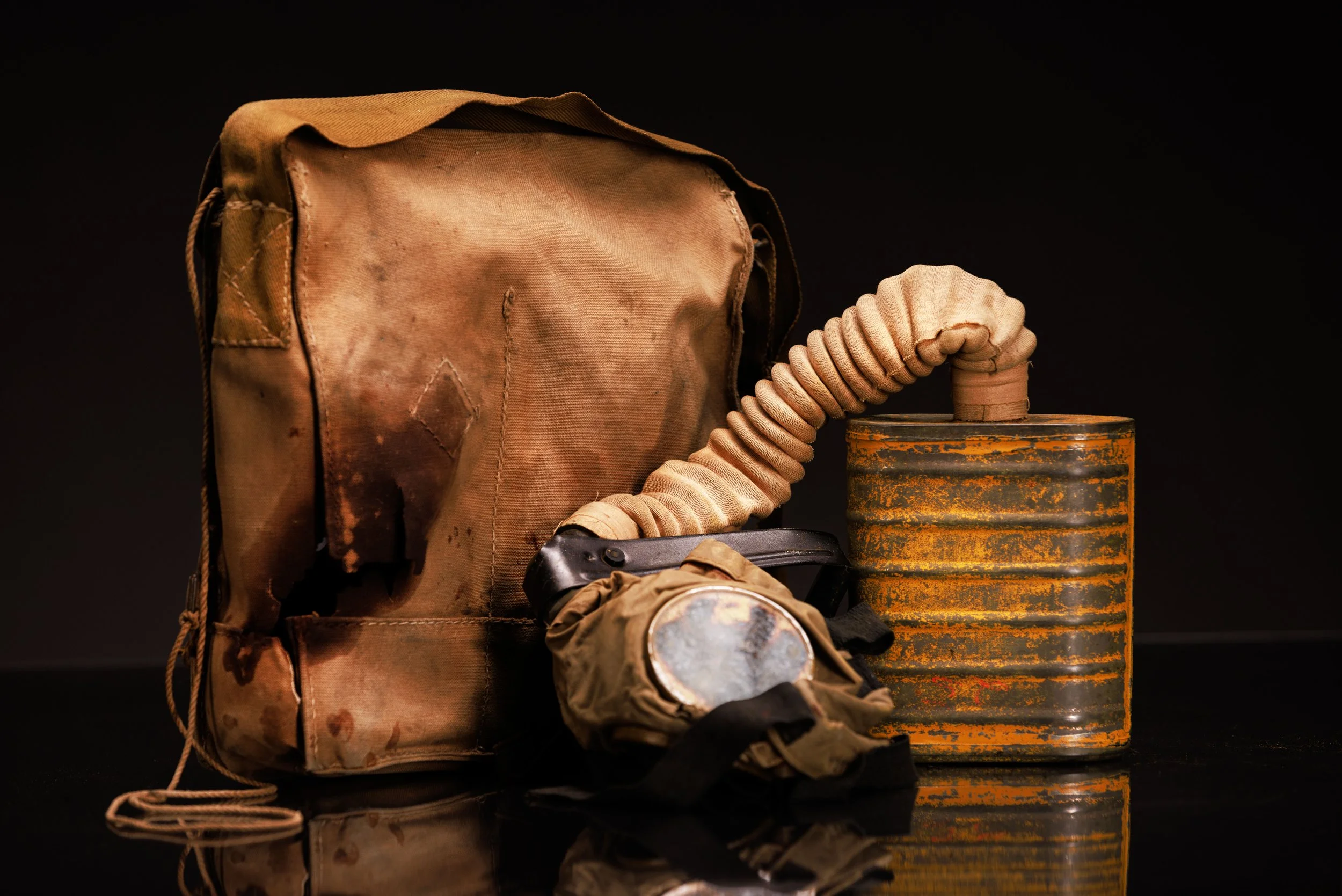 blood-stained military backpack with a coiled hose attached to a rusted metal canister, set against a black background.