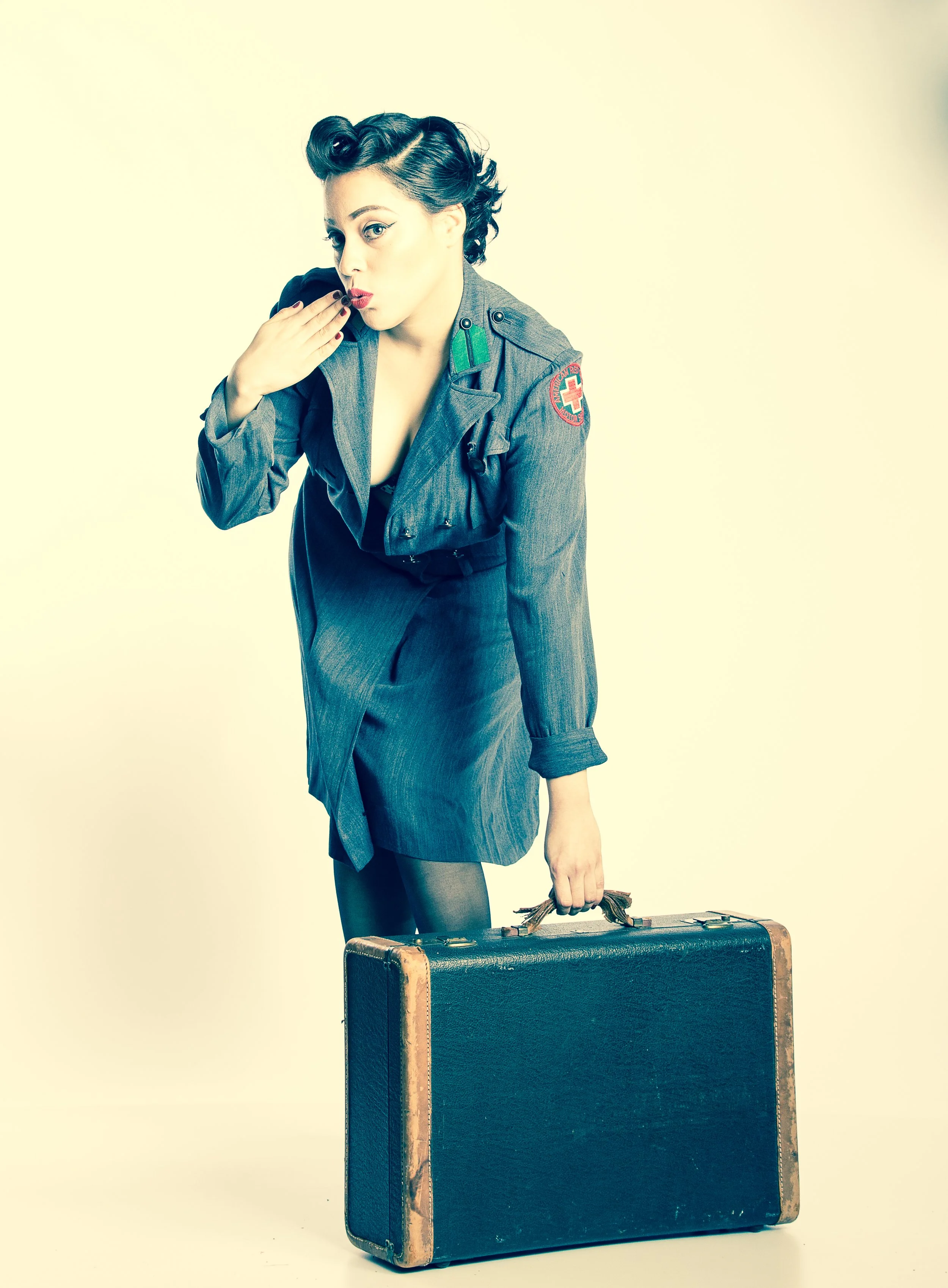 A woman dressed in a vintage military nurse uniform, holding a suitcase, and blowing a kiss.
