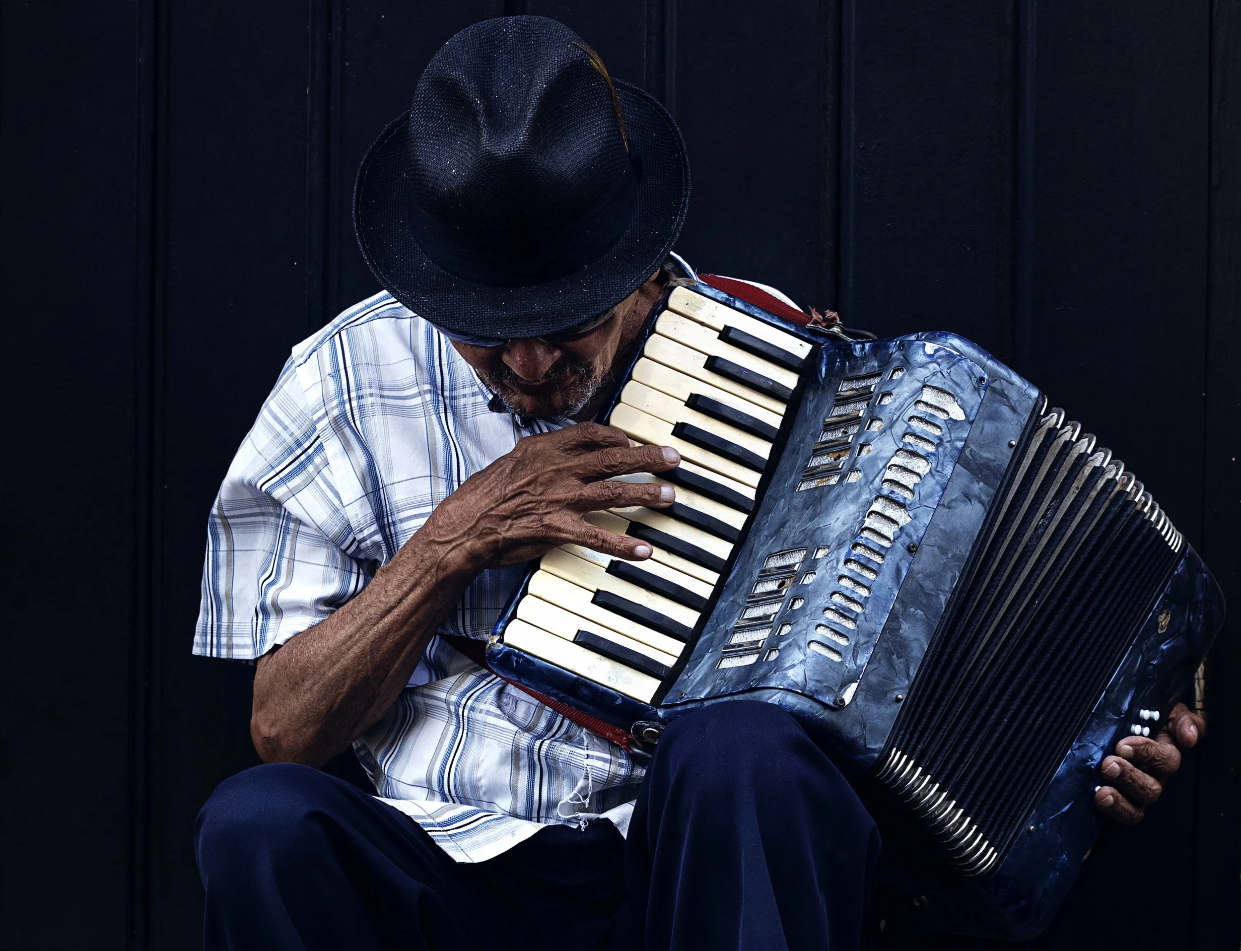 An older man wearing a striped shirt and dark hat playing an accordion against a dark background.