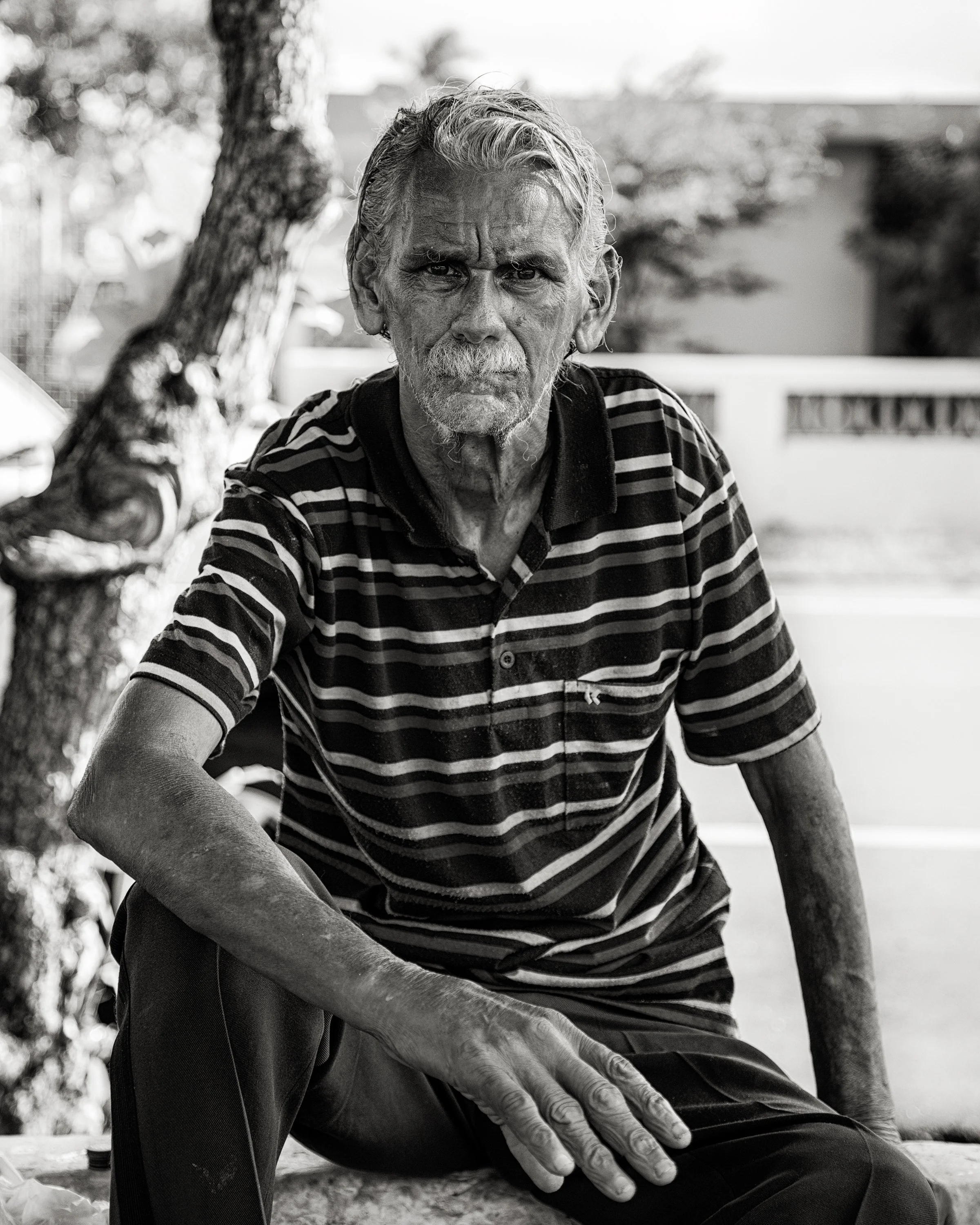 An elderly man with gray hair, a mustache, and a beard, sitting outdoors near a tree, wearing a striped shirt, with a serious expression on his face.