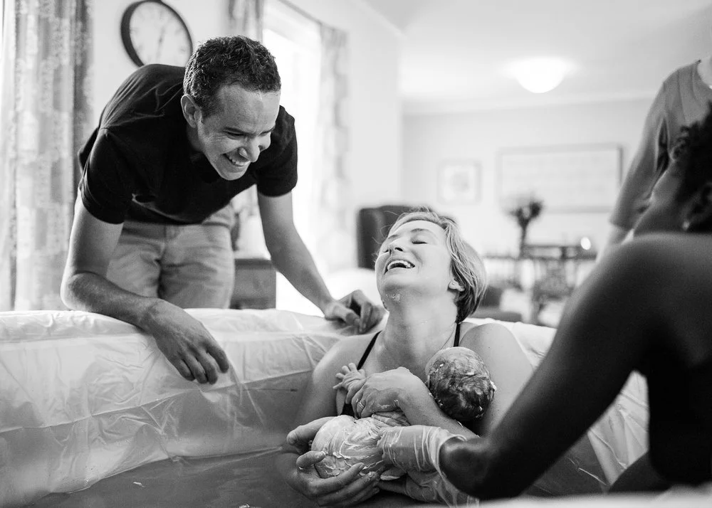 smiling woman in a birth tub who has just delivered her baby at home with her husband and midwife looking on