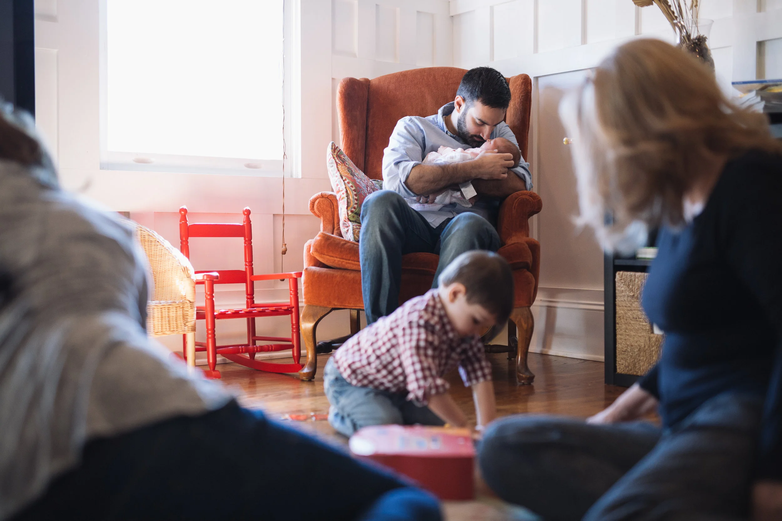 Dad snuggling with newborn baby while mom and brother play on the floor at home