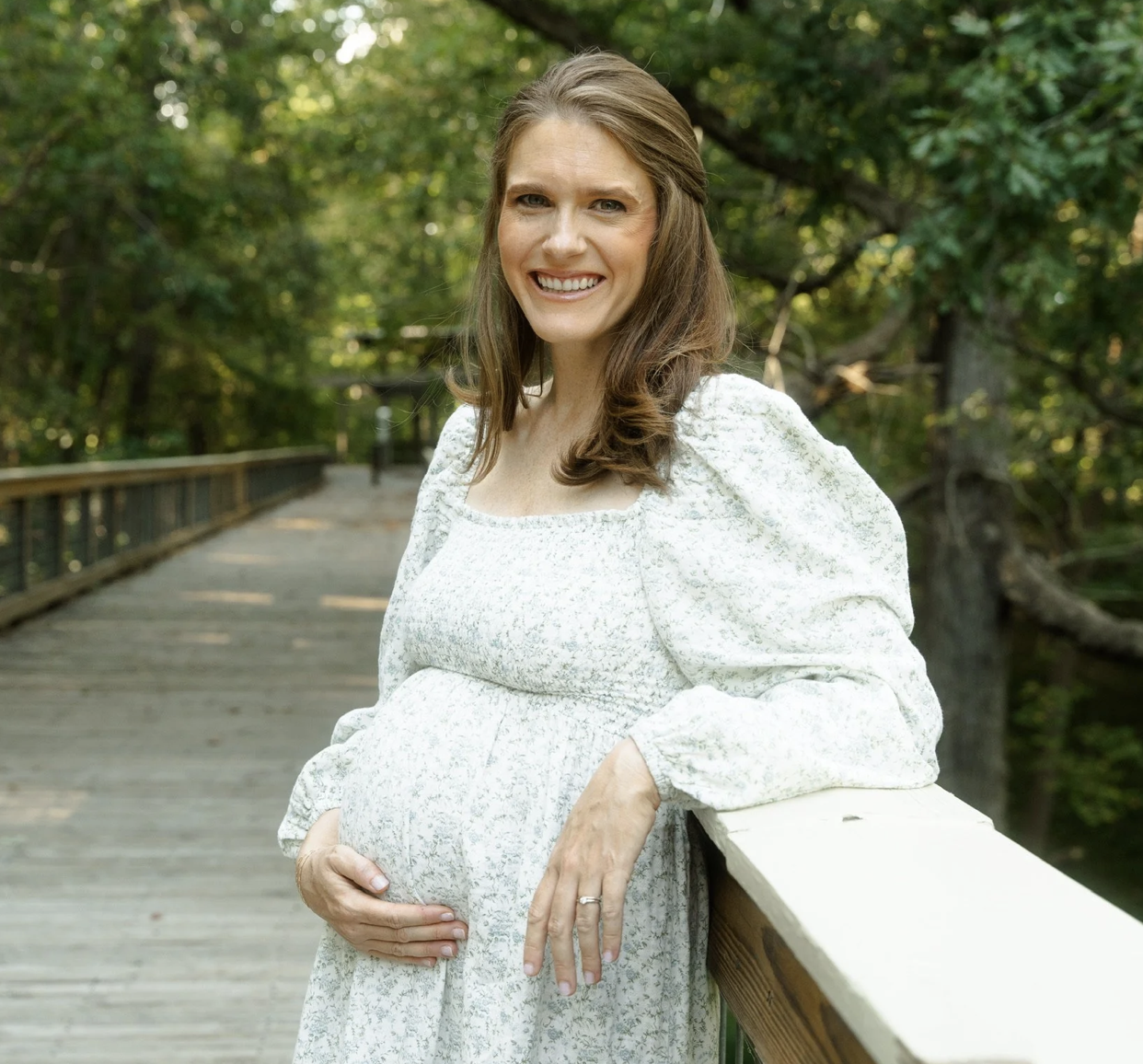A pregnant woman with long brown hair smiling while standing outdoors on a wooden bridge surrounded by green trees.