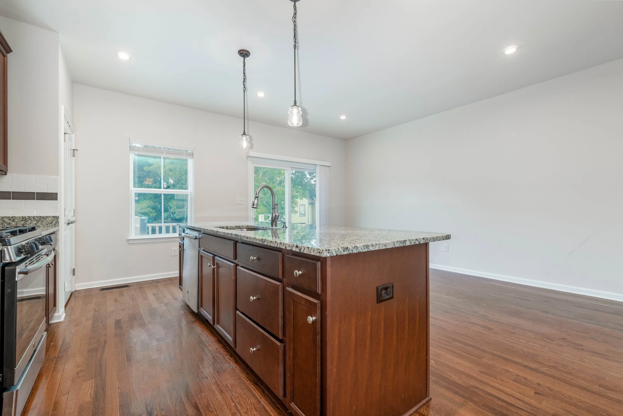Modern kitchen interior with granite countertop island, hardwood floors, stainless steel appliances, pendant lighting, and large windows.