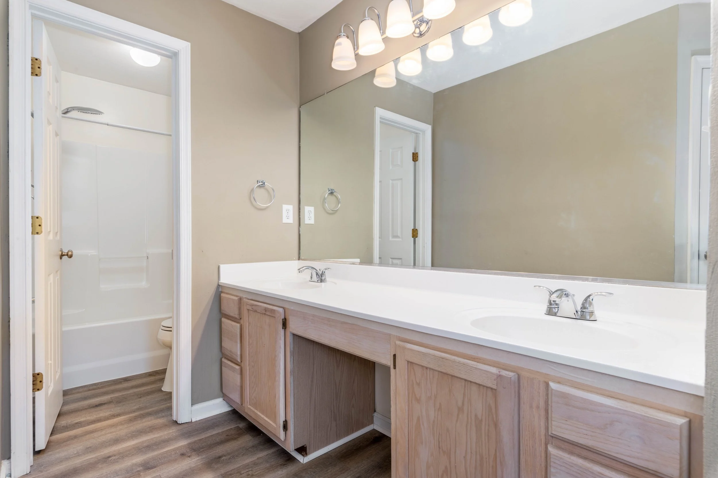 Bathroom interior with wooden vanity, dual sinks, large mirror, overhead lighting, and visible shower area.