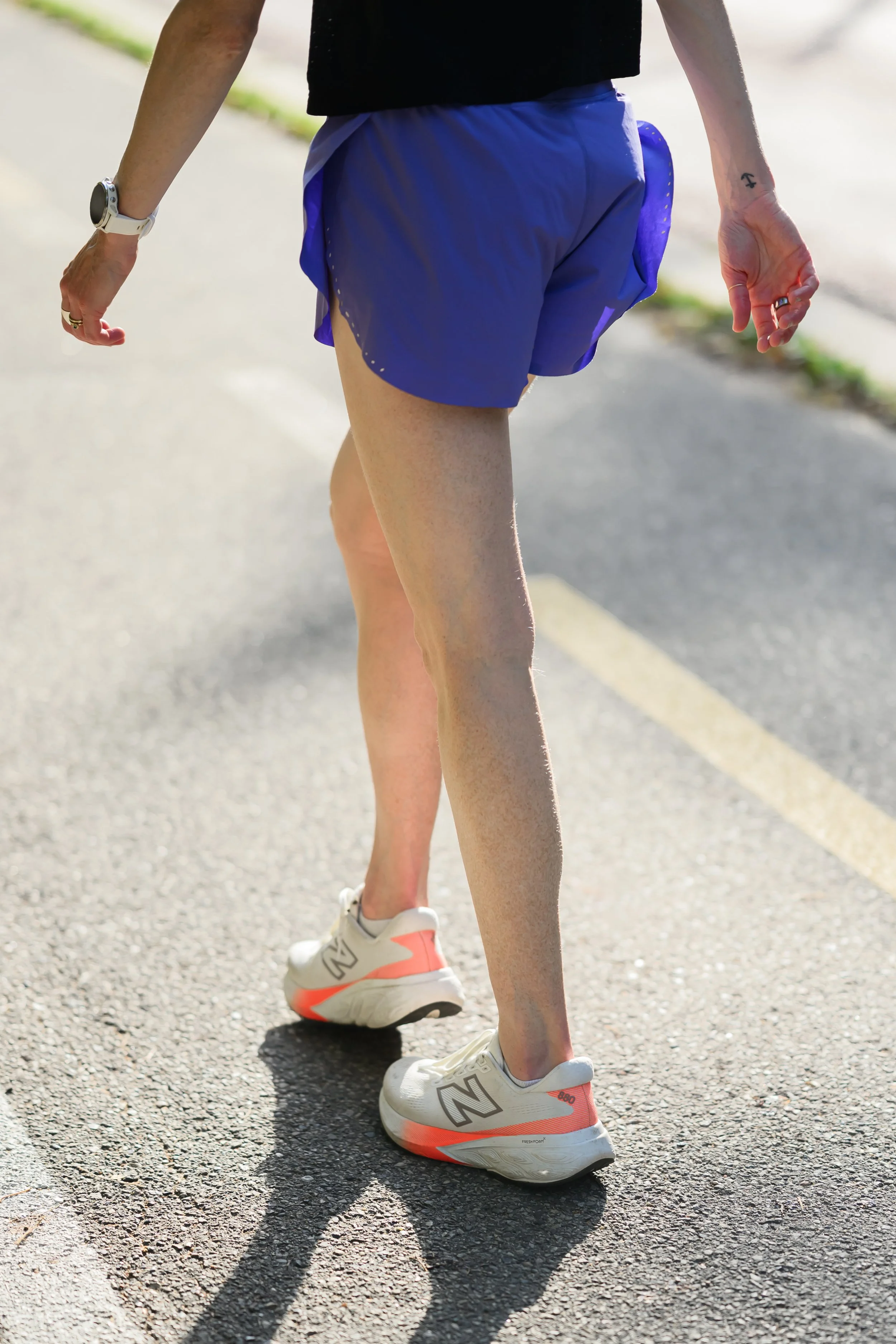 Person walking on a paved road, wearing blue shorts, white New Balance running shoes, a black top, a white smartwatch, and a ring.