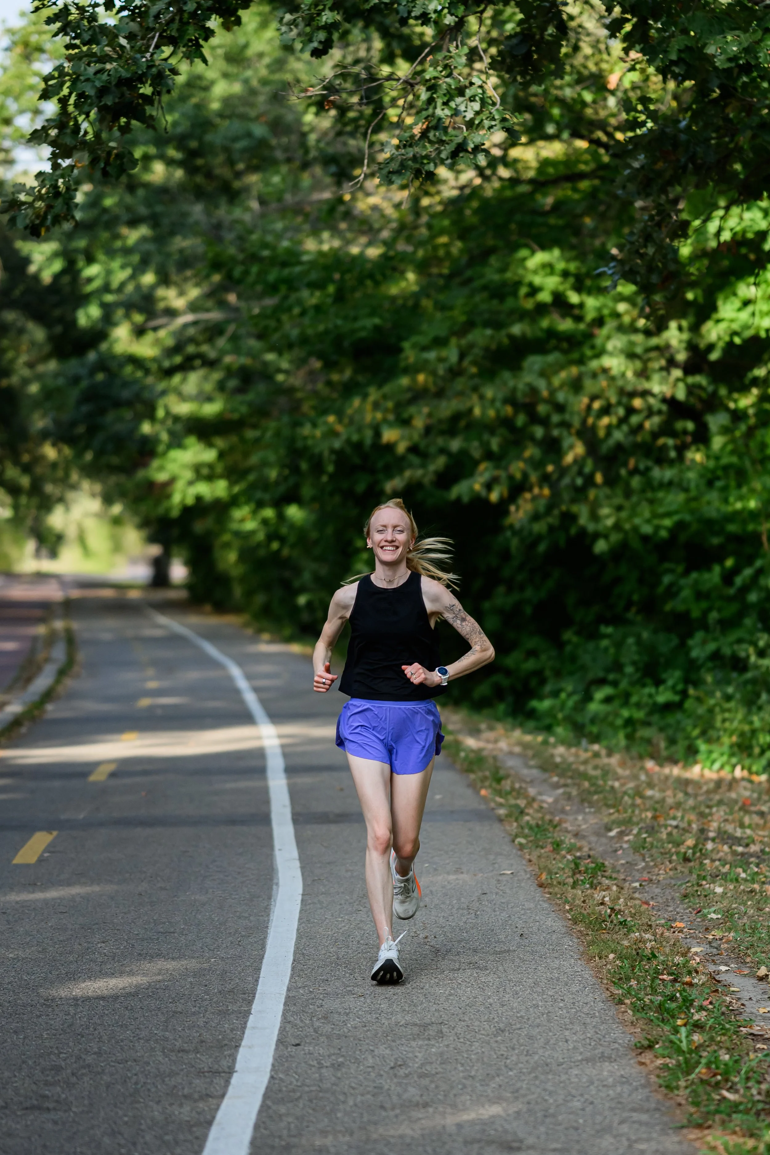 A woman is running outdoors on a paved path lined with trees, smiling and wearing athletic clothing, including a black sleeveless top, purple shorts, and running shoes.
