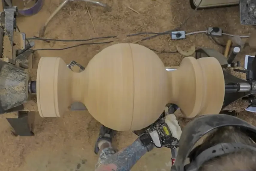 Woodturning lathe with a large, unfinished wooden vase or urn shaped object in the process of being crafted.