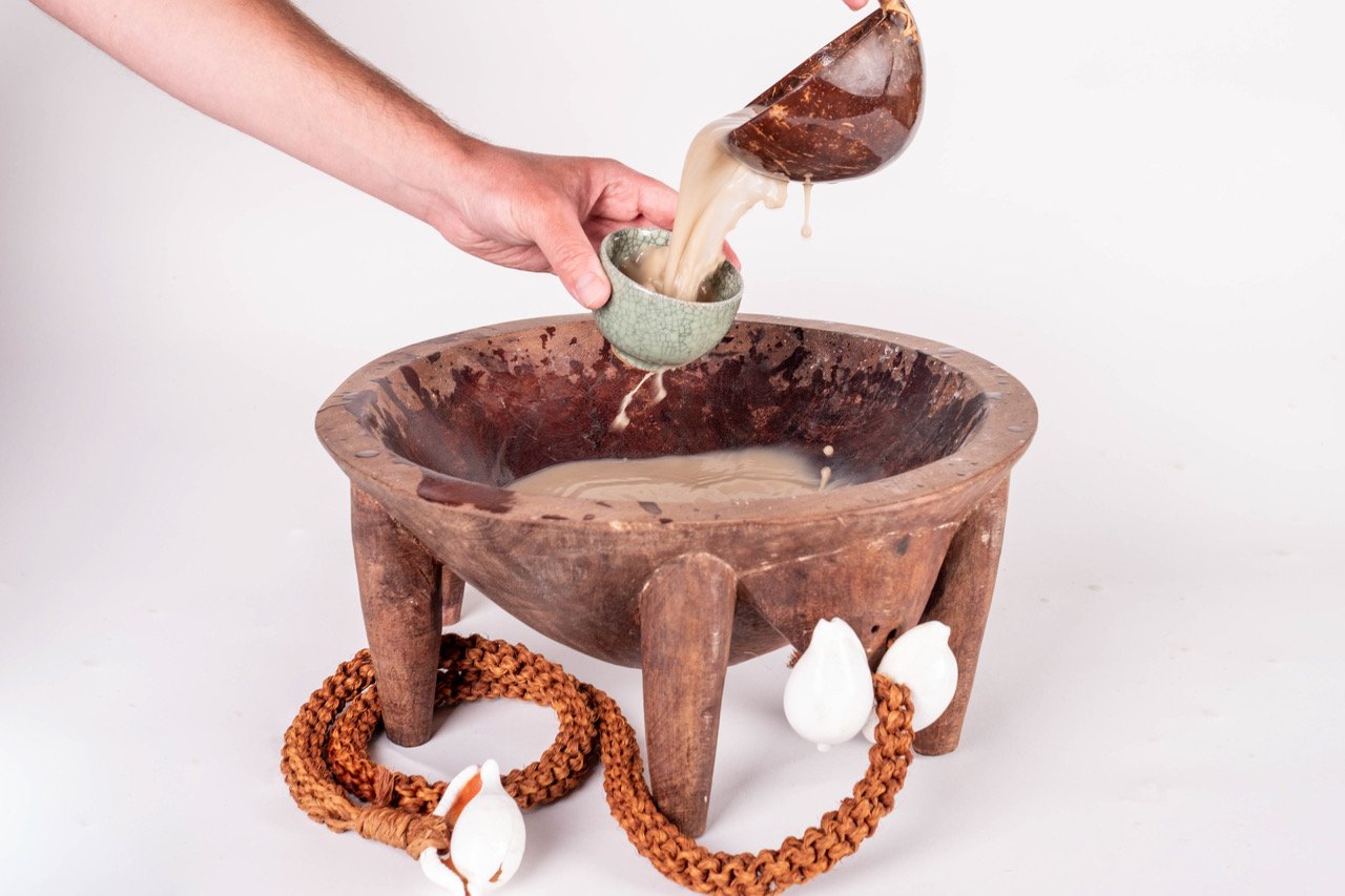 Hand pouring liquid into a small cup from a traditional wooden bowl with three legs. The bowl has a decorative cord with white ornaments.