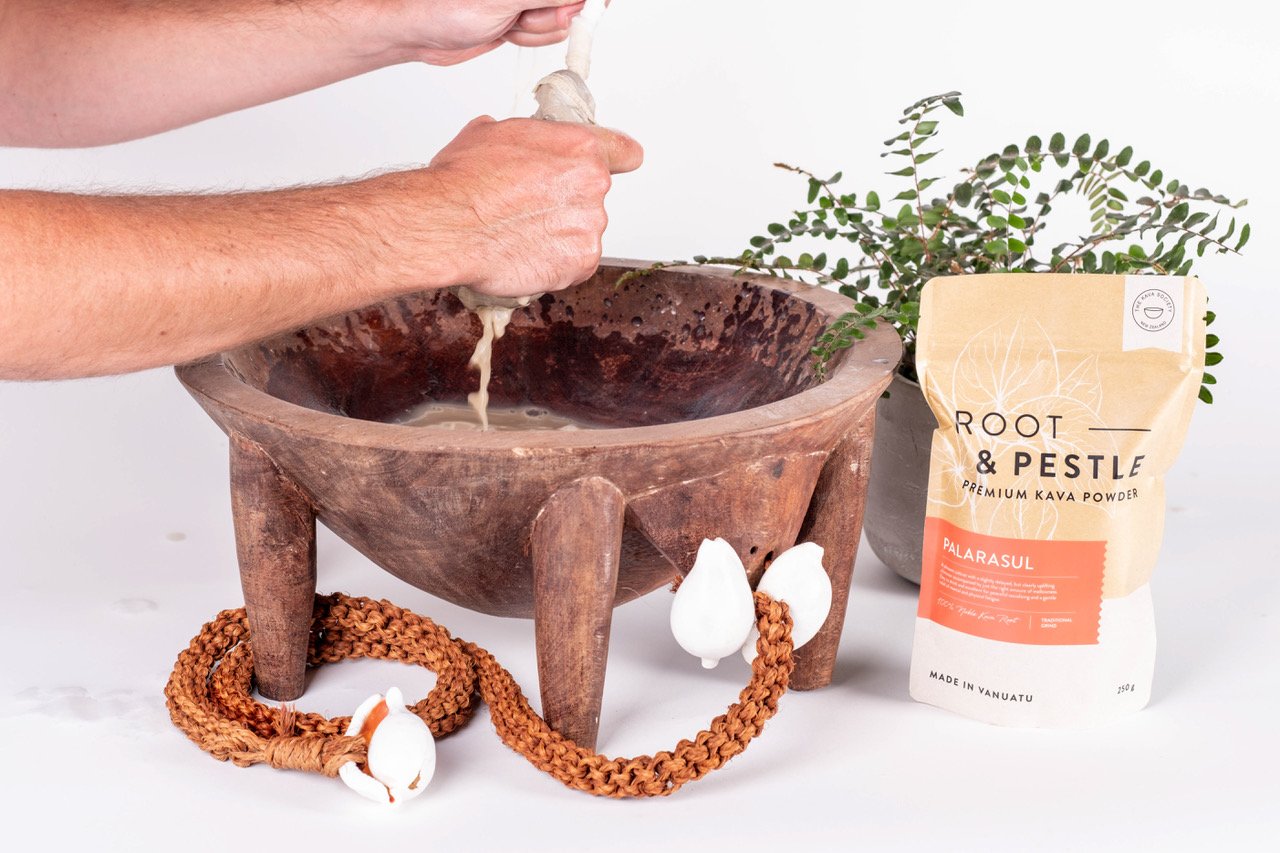 Hands preparing kava drink by squeezing liquid into a wooden bowl, with a package of Root & Pestle premium kava powder nearby, a fern plant, and a rope with white objects.