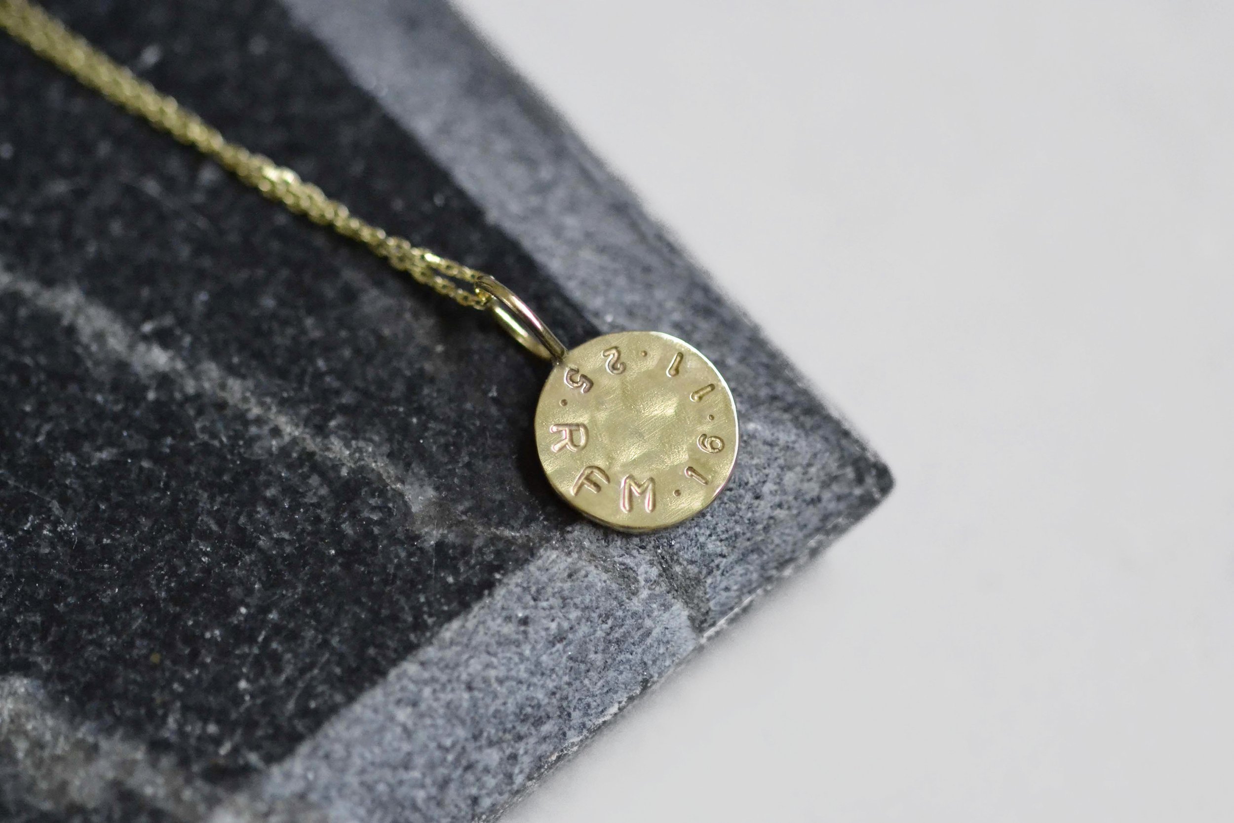 A gold-toned necklace with a small round pendant engraved with letters and numbers, resting on a dark textured surface.