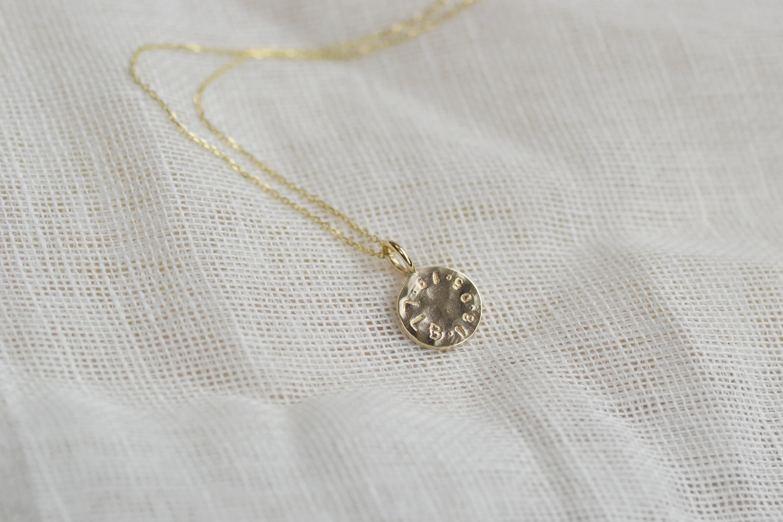 A delicate gold-toned necklace with a small round engraved pendant, displayed on a white textured fabric background.