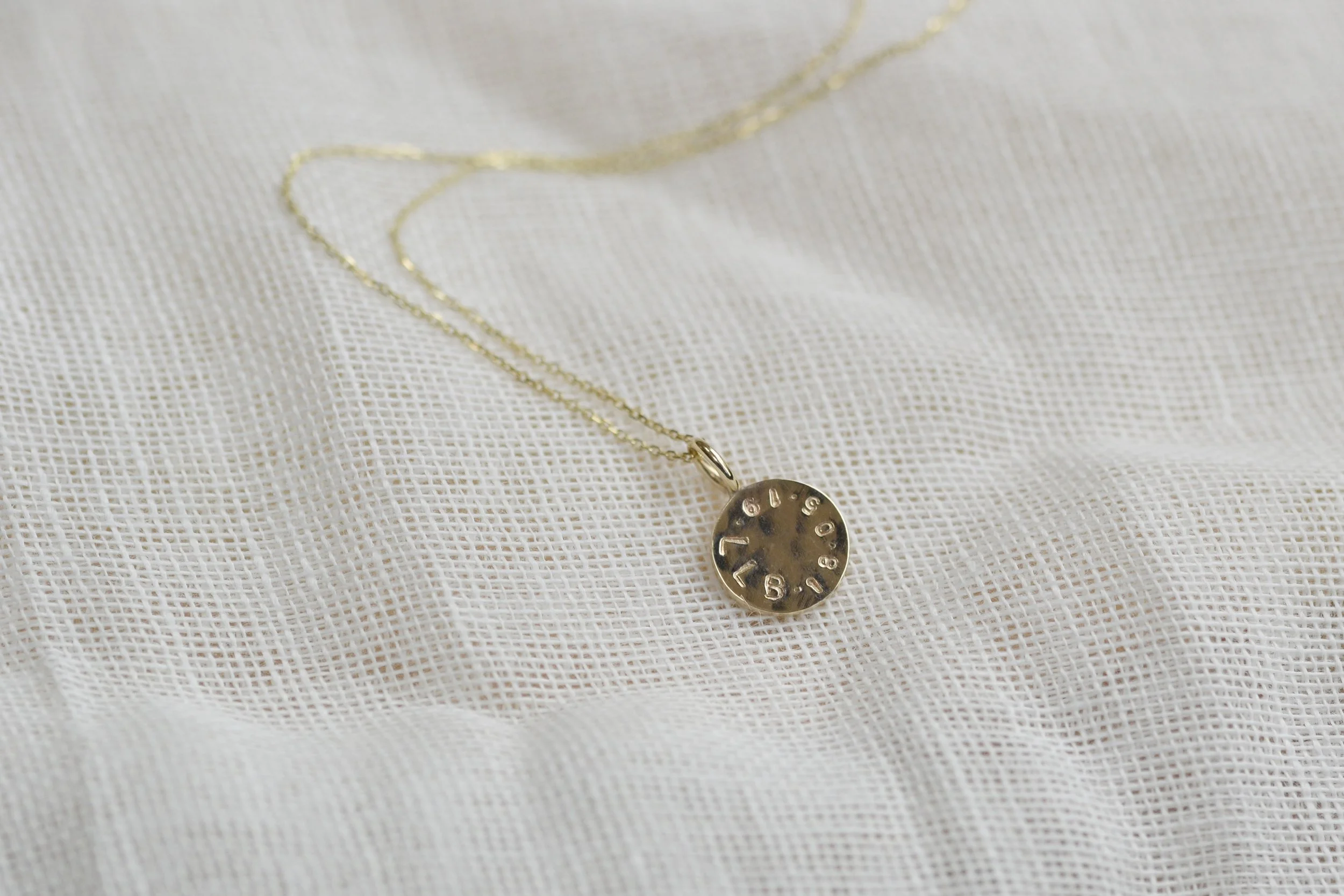 A delicate gold-toned necklace with a small round engraved pendant, displayed on a white textured fabric background.