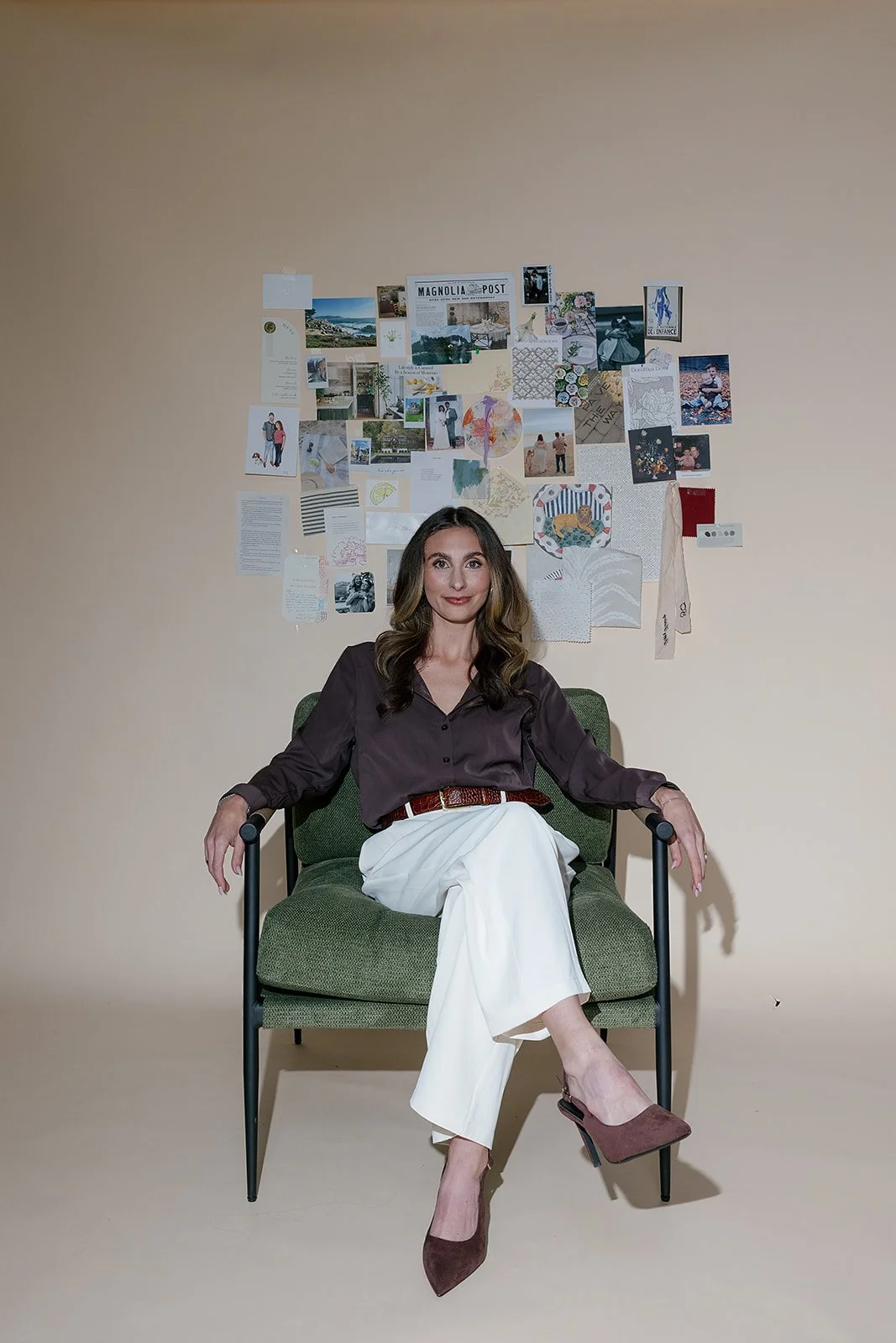 A woman with wavy brunette hair sitting in a green armchair against a beige wall with a collage of photos, artwork, and notes.