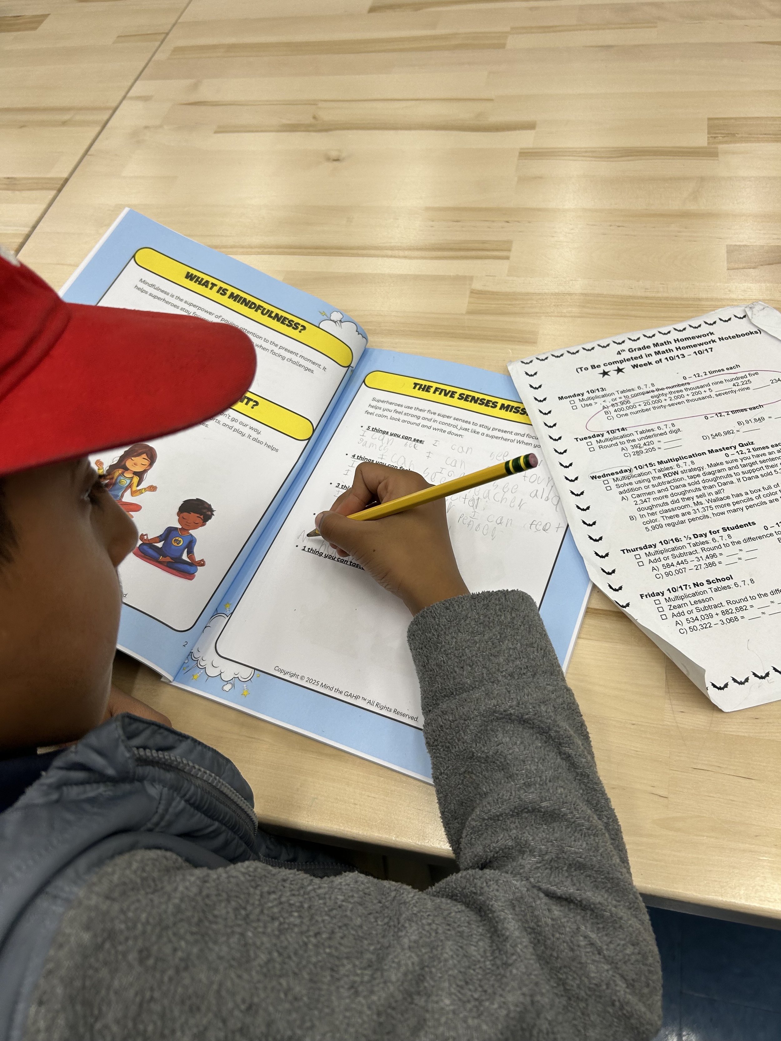 A child wearing a red hat and a gray jacket seated at a wooden table, writing in a notebook with a yellow pencil. There are school papers and worksheets on the table, including a math homework sheet and a science-themed booklet titled 'What is Mindfu