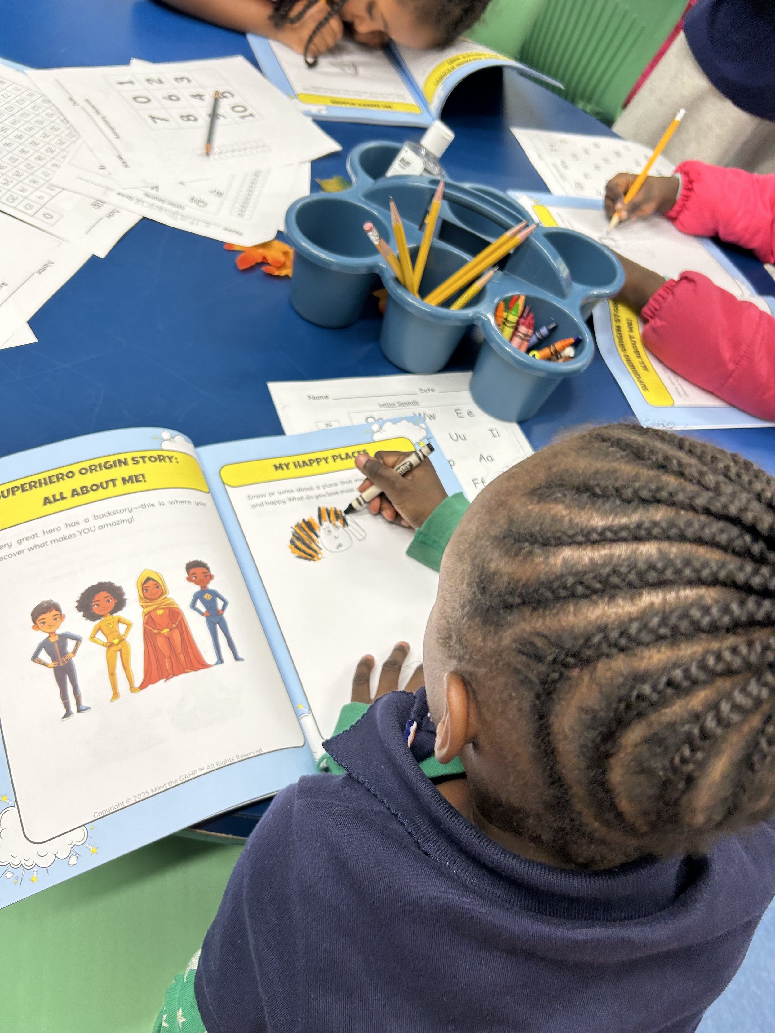 A young refugee elementary school age child at a table, working on the Mindful Superheroes activity book with character illustrations and writing activities, surrounded by school supplies, with other children visible nearby.