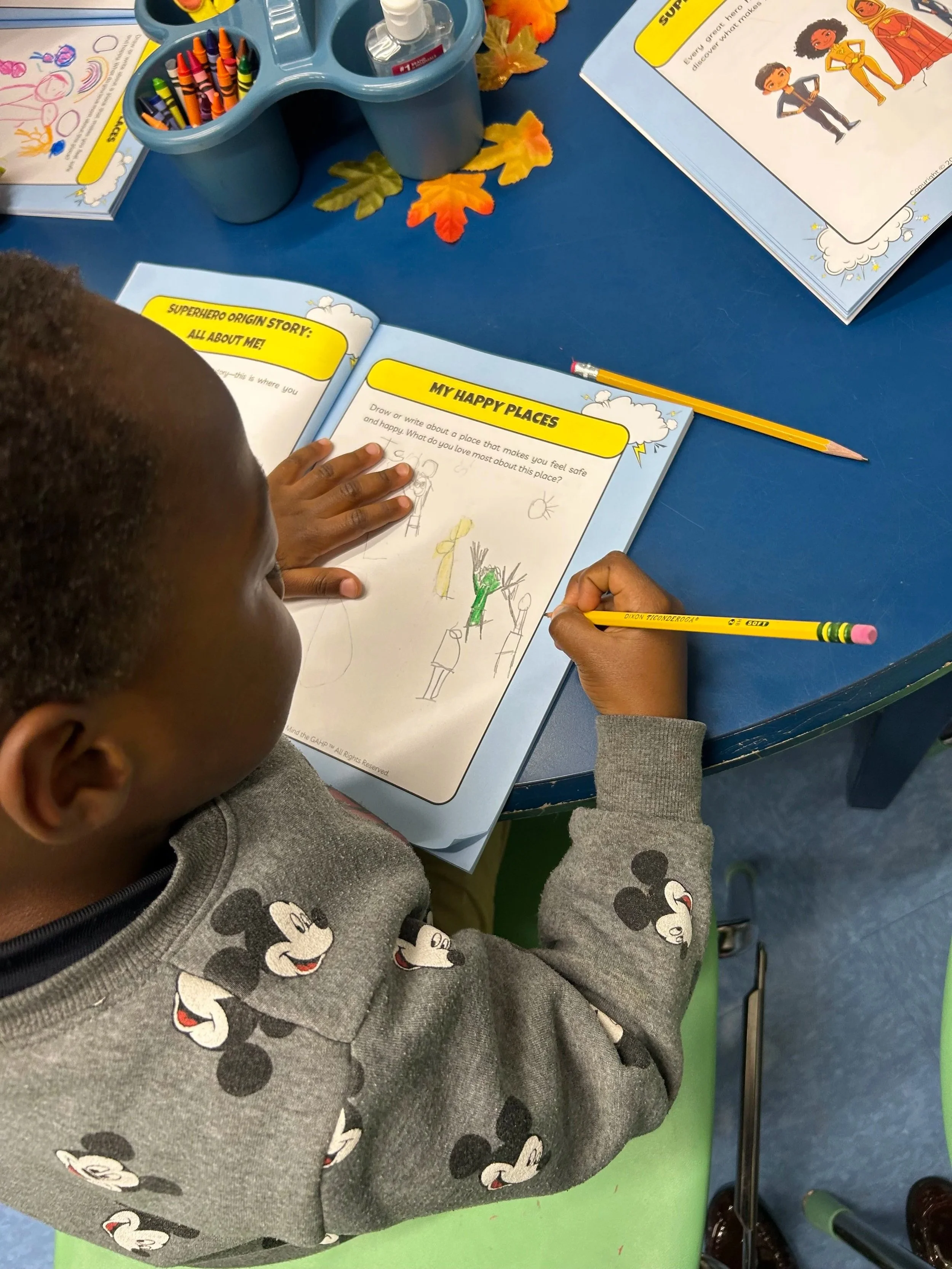 A young child is sitting at a blue table working on a drawing activity in a children's workbook. The child is wearing a gray Mickey Mouse sweater and has short hair. On the table, there is a yellow pencil, a blue plastic container with crayons, and a