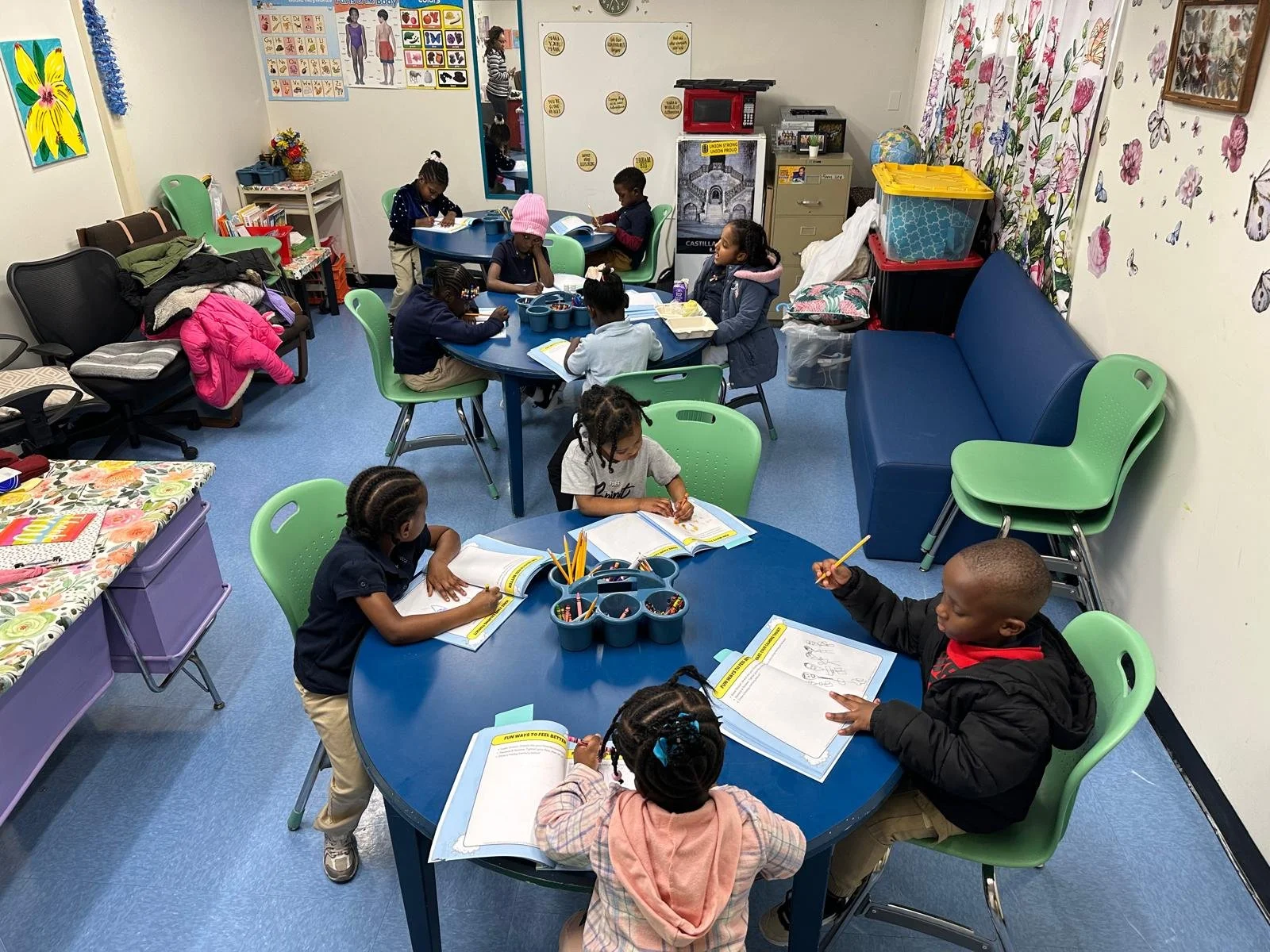 A classroom with young children sitting at blue and green tables, engaged in reading and writing activities. The room has colorful decorations, posters, and storage containers.