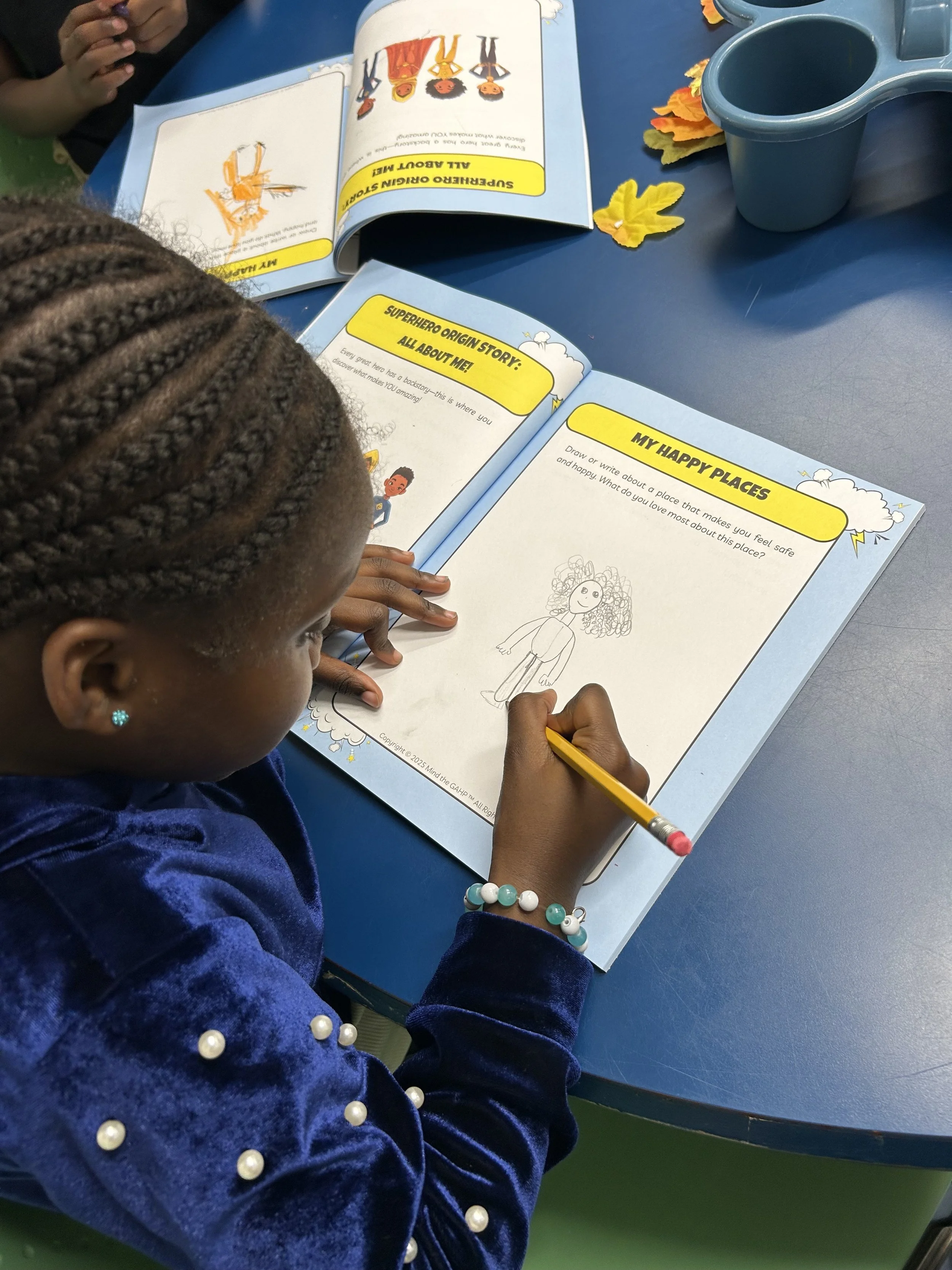 A young girl with braided hair and a pearl bracelet draws on an open workbook at a blue table, with autumn leaf decorations nearby.