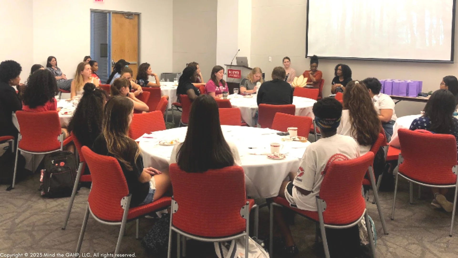 A group of young women and girls at a university, listening to a presentation or speaker at the front of the room. There is a large screen on the wall behind the speaker, and purple gift bags on a table to the right.