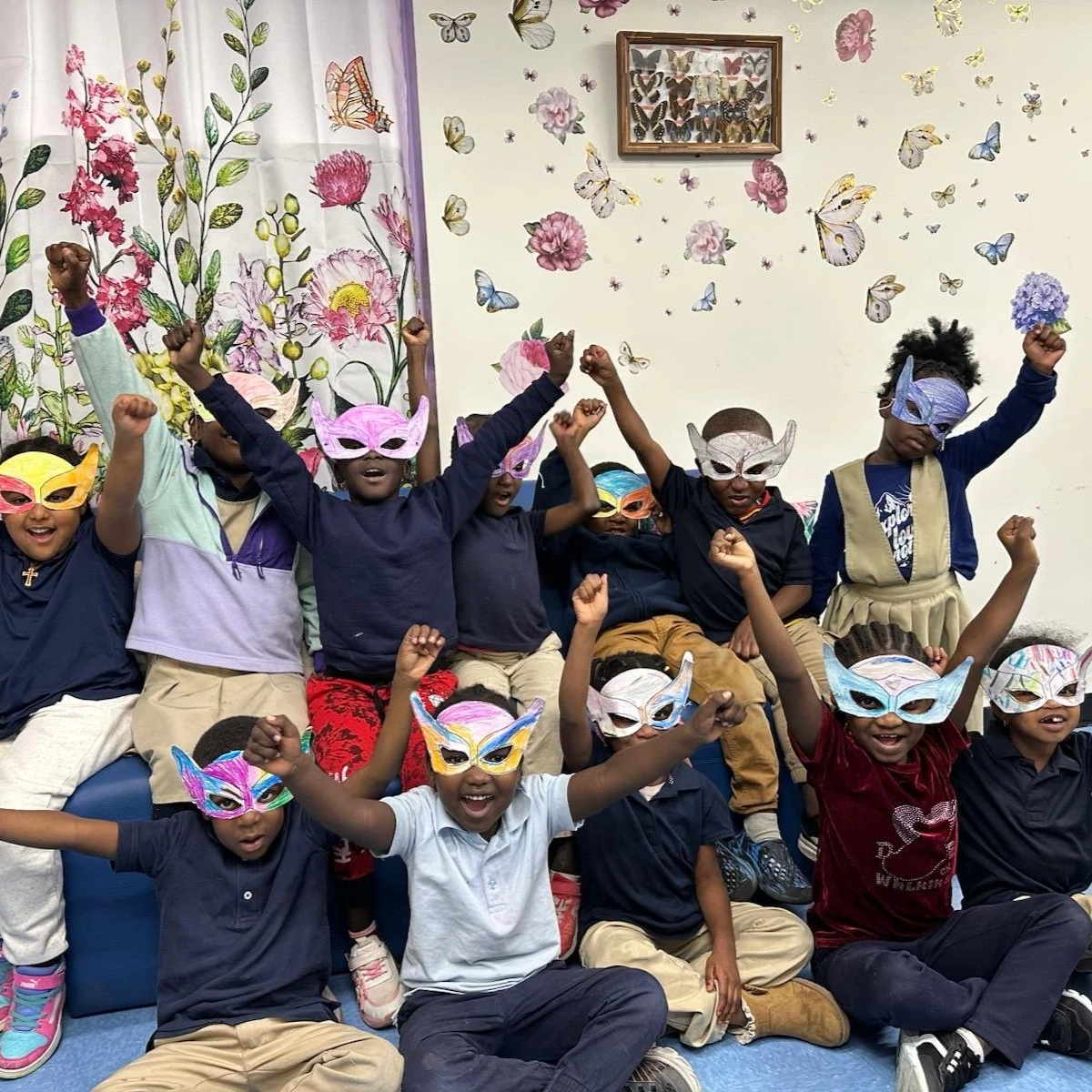 A group of children wearing colorful masks and celebrating, posing with raised fists in a decorated room with butterfly and floral wallpaper.