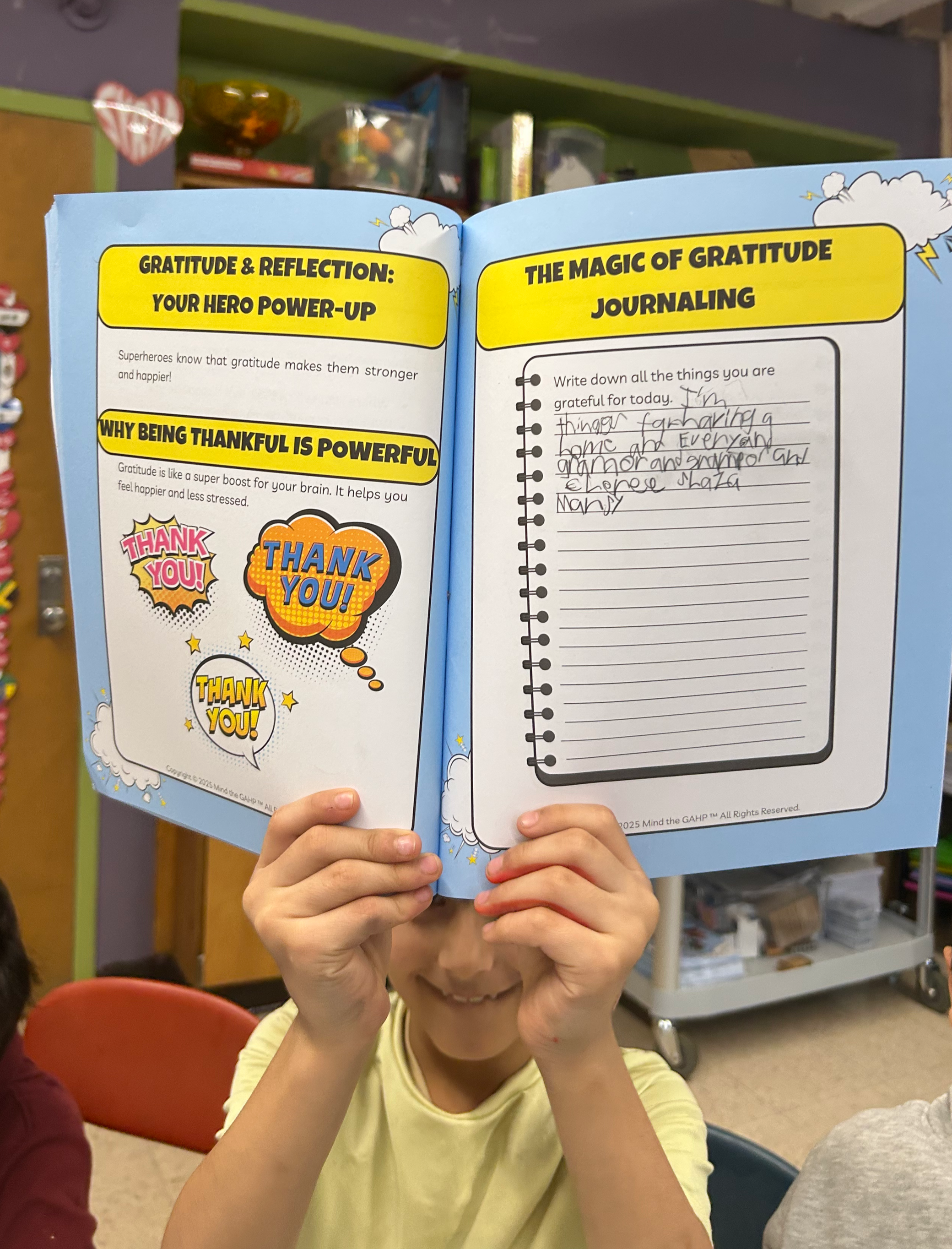 Child holding an open workbook in front of their face with hands at the bottom of the image, focus on the workbook which has colorful graphics and a handwritten note inside.