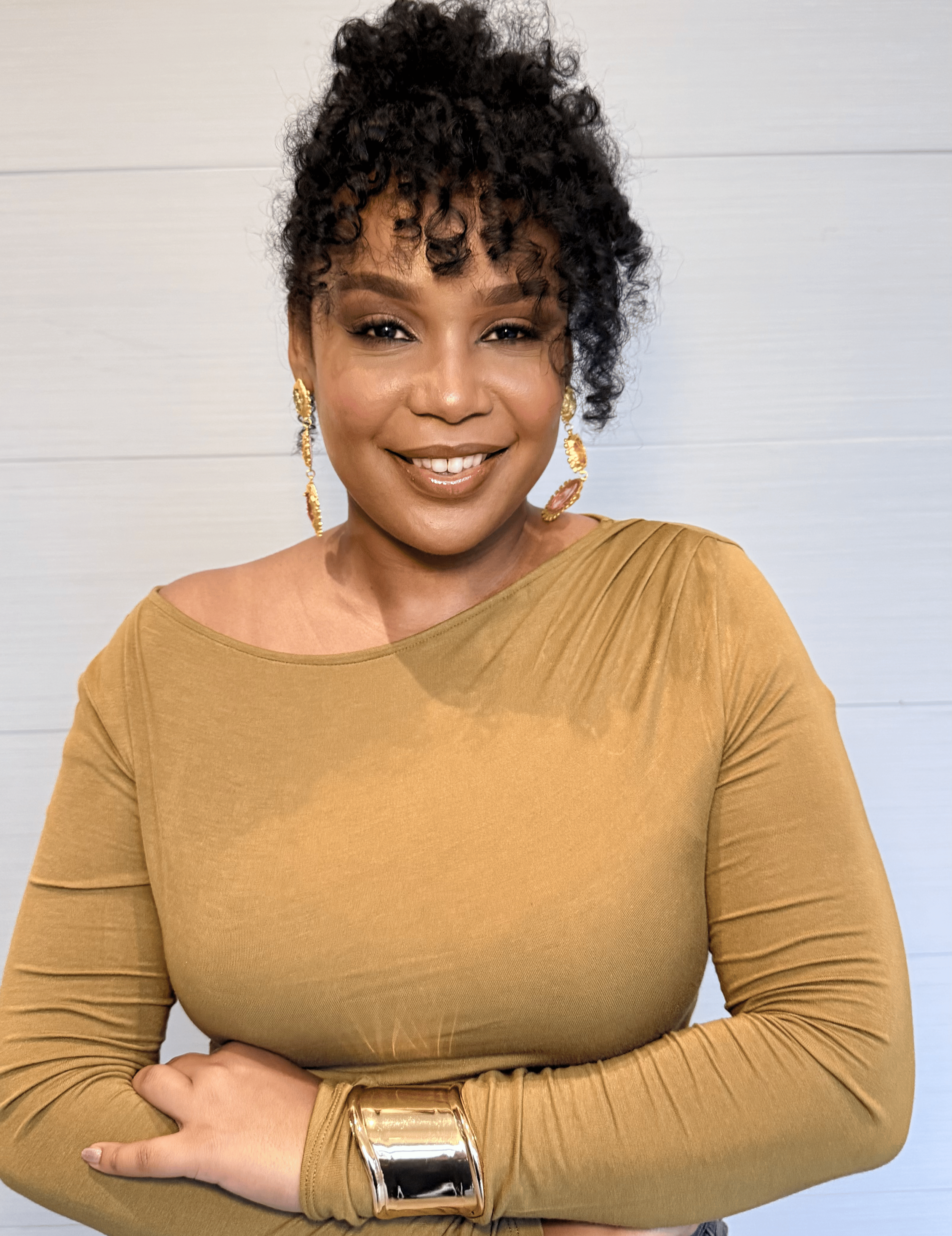 A woman with dark, curly hair in an updo, wearing large gold earrings and a gold dress with long sleeves, standing against a light-colored background.