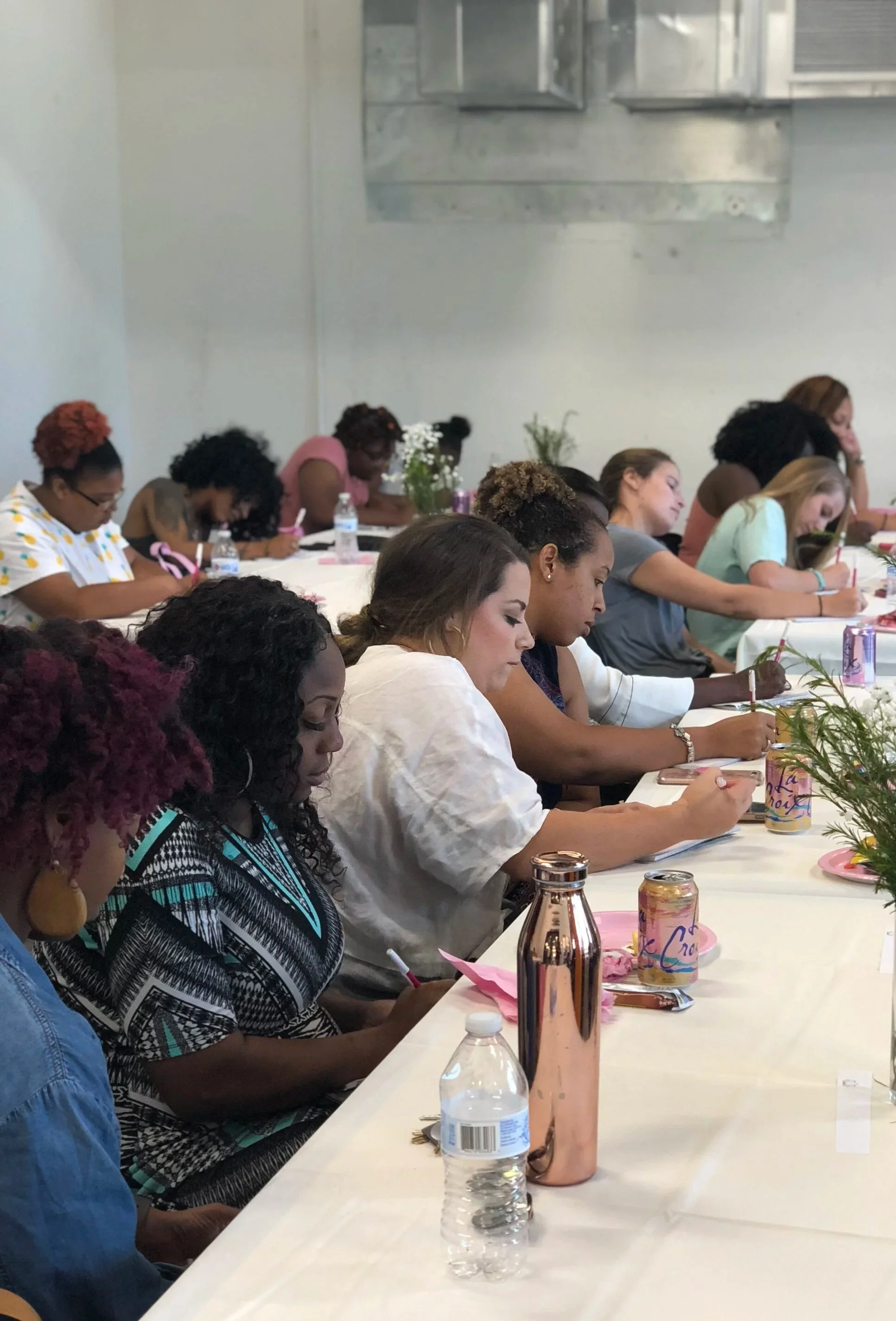 A group of women sitting at a long table, engaged in a work-life balance workshop with various drinks and decorations on the table, in a bright indoor setting.