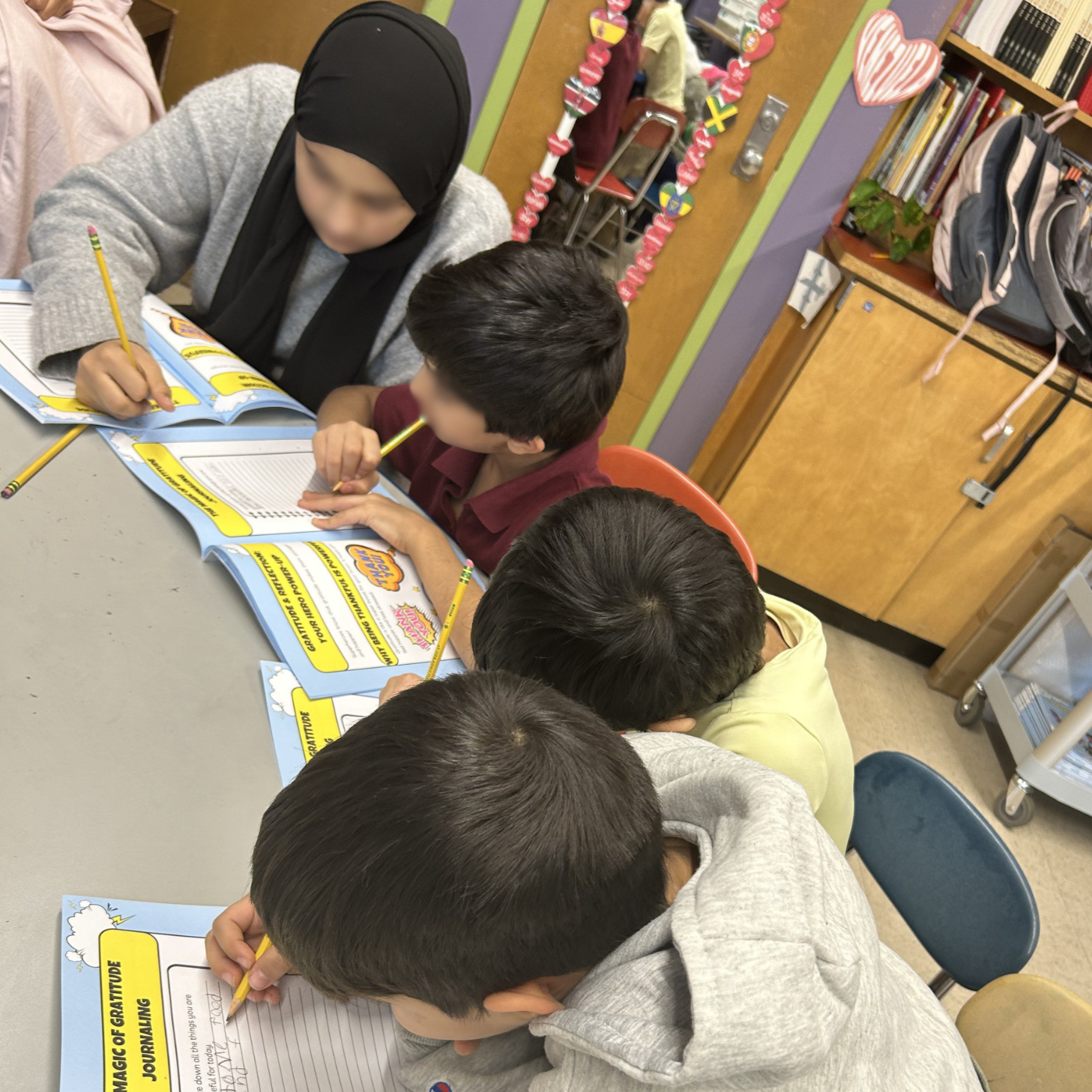 Children sitting at a table in a classroom writing in journals with yellow pencils, with bookshelves and colorful decorations in the background.