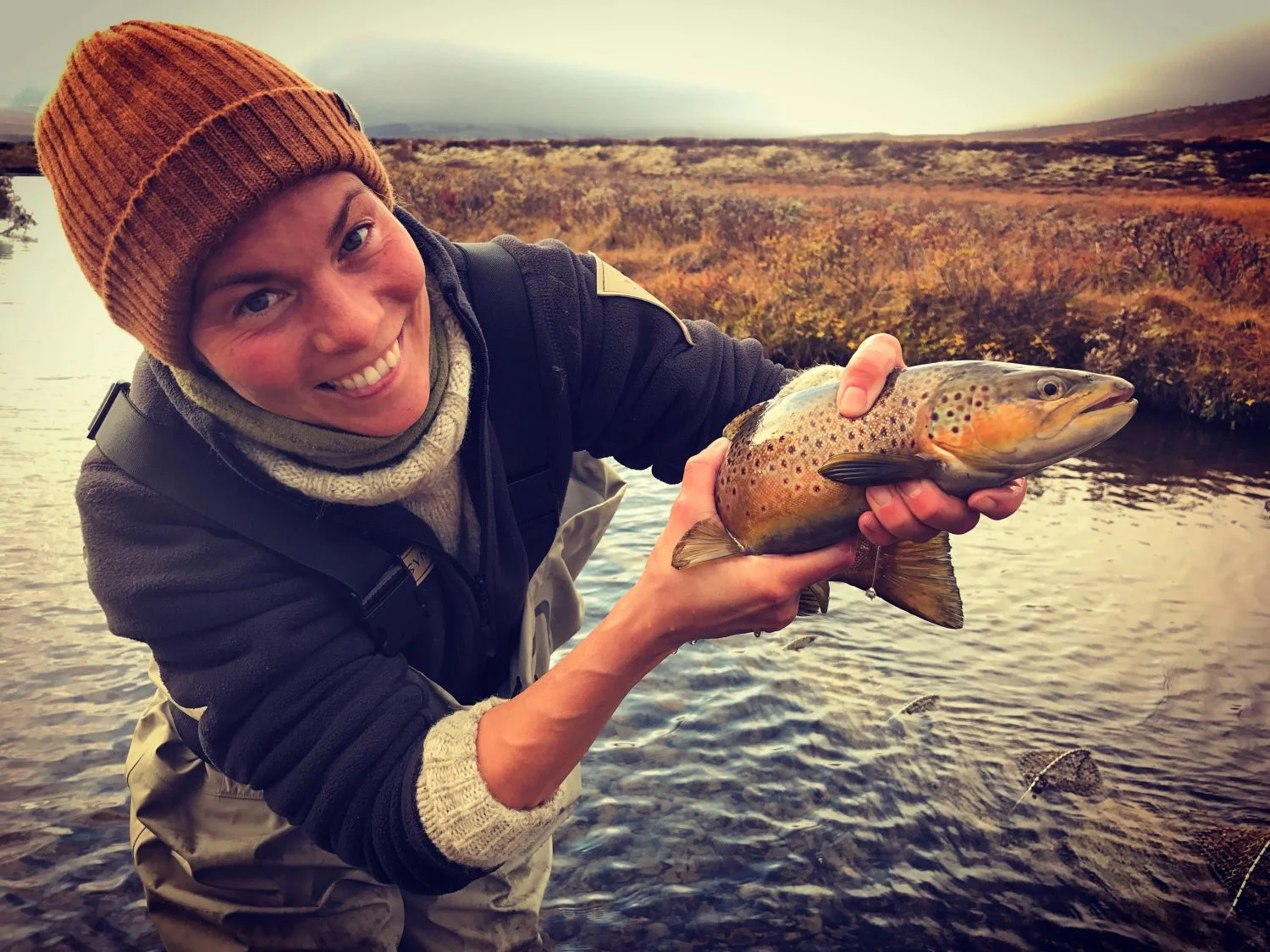 En person smilende som holder en fisk med begge hender ved en elv eller innsjø, omgitt av høstfarger.
