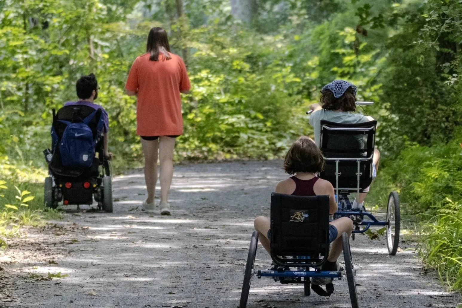 A group of people enjoy a even stone dust trail surrounded by greenery. One person is in a motorized wheelchair, one is walking, and two are using adaptive bicycles.