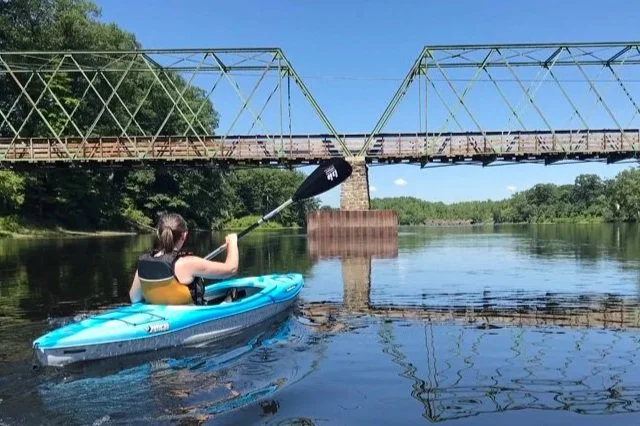 A woman wearing a yellow life jacket in a blue kayak paddles towards a large steel span bridge that goes across a river. There are trees lining the shore and a blue sky. The bridge, trees, and sky are reflected in the calm water.