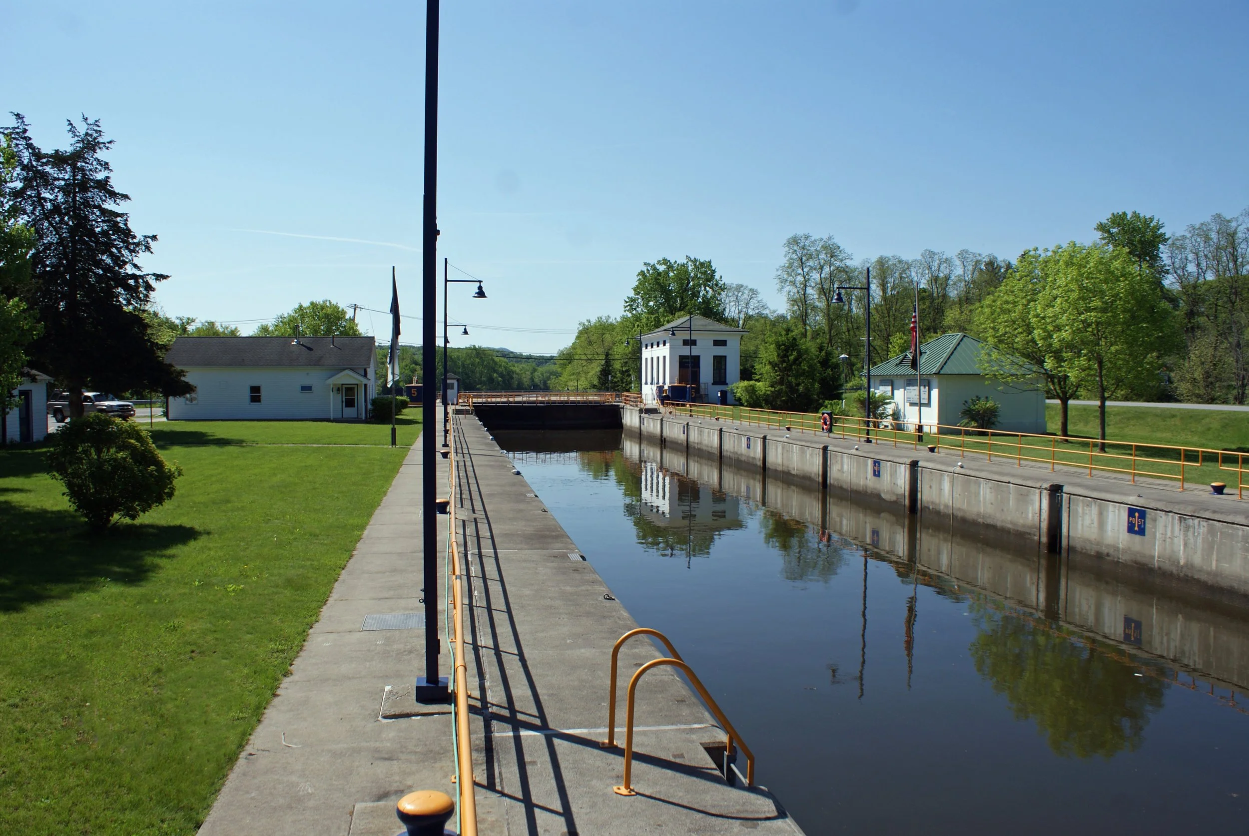 A photo of Lock C5. The lock is made up of cement walls with a channel of water going through the center. The lock is surrounded by green grass. White lock tending buildings are in the background, and the sky is a brilliant blue.